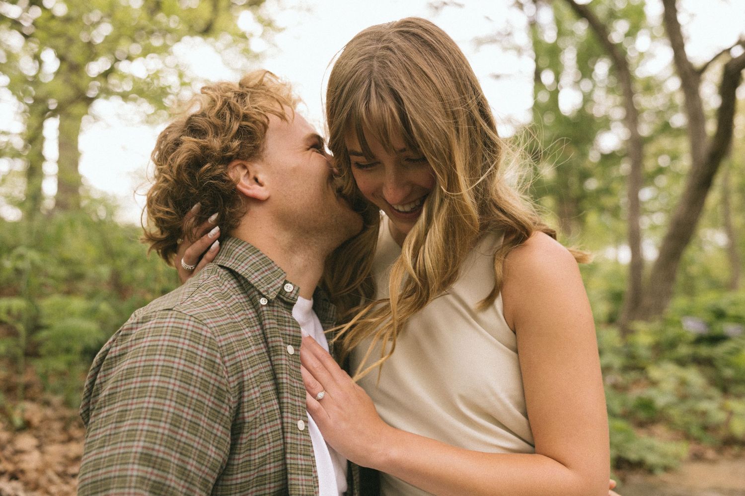 A romantic moment between a couple in casual attire sharing an intimate moment outdoors in a wooded setting.