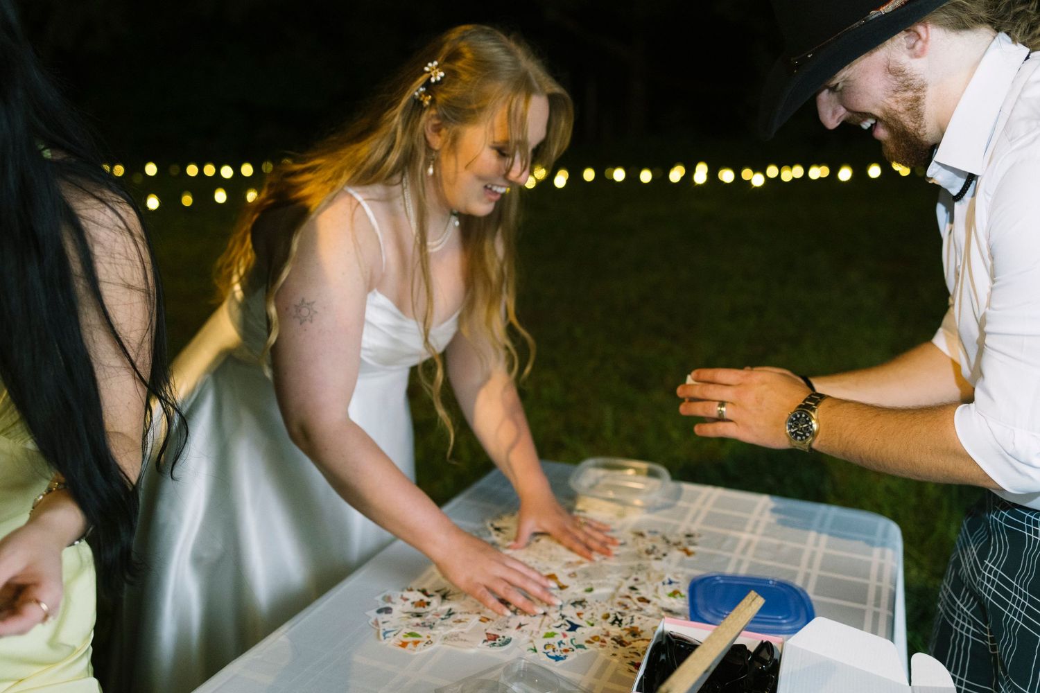 Someone in a white dress plays dominoes on an outdoor table at night with string lights in the background.