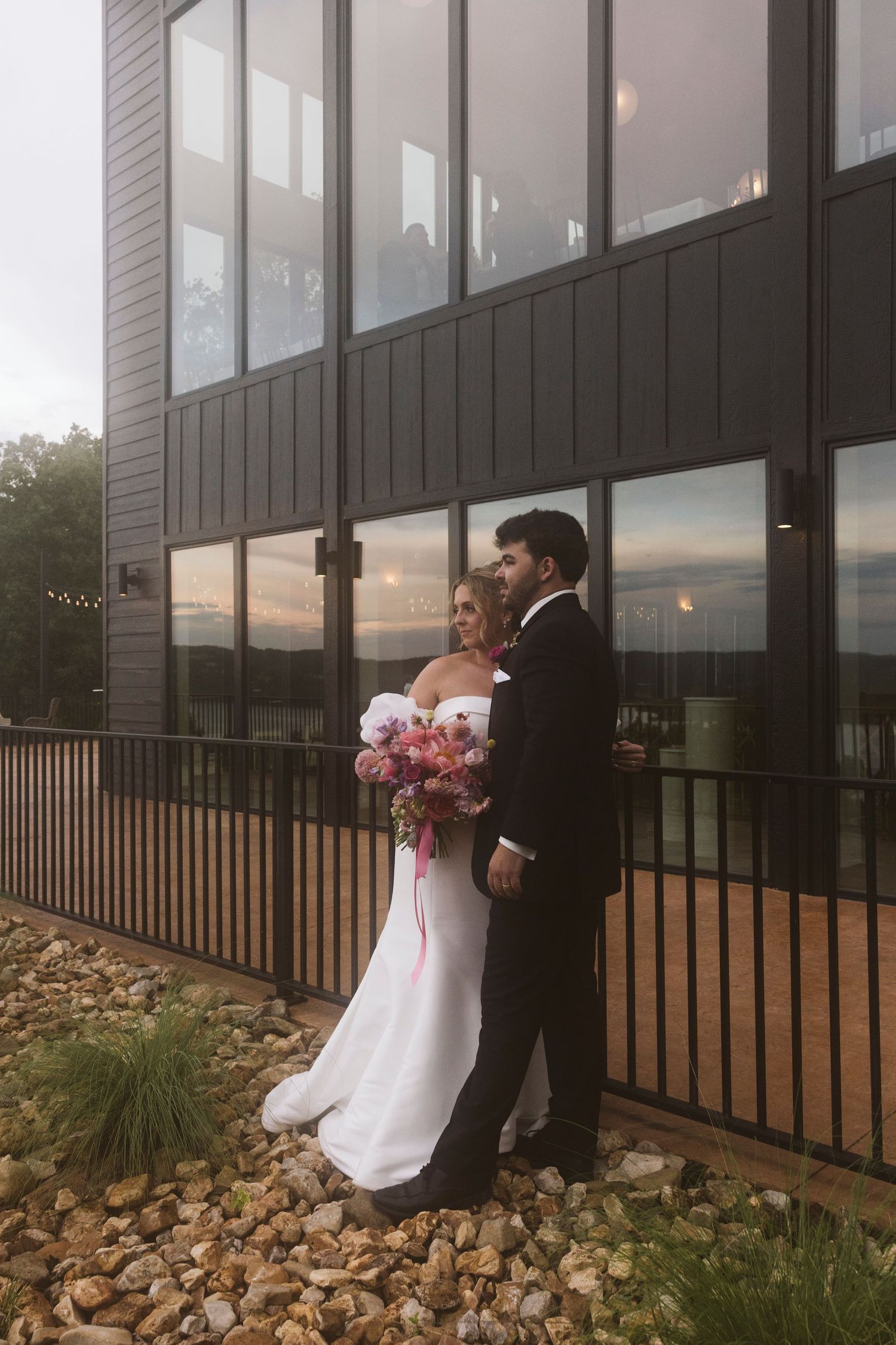 A couple in wedding attire stands outside a modern building with floor to ceiling windows at sunset.