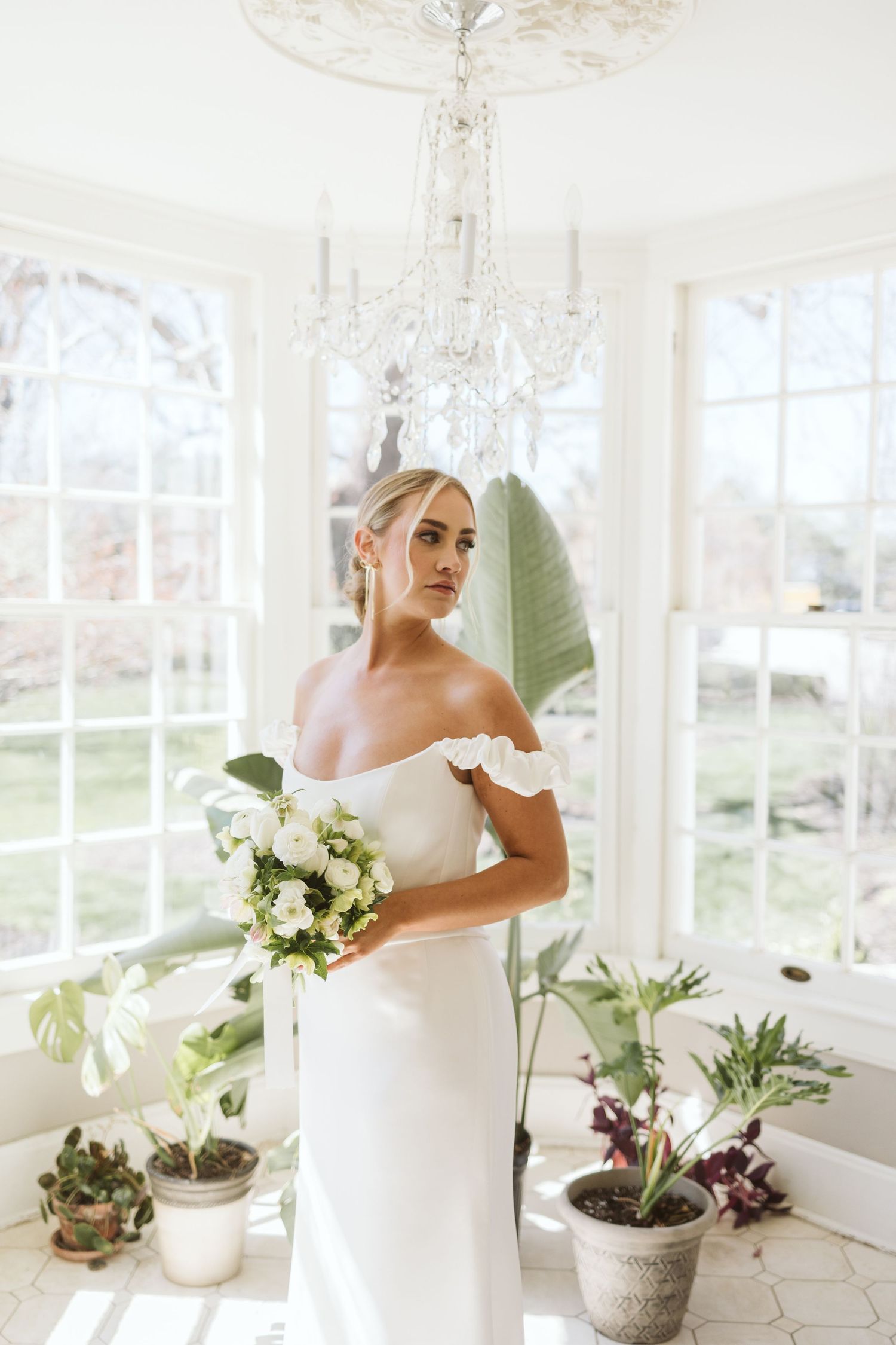 An elegant off-shoulder white wedding dress and bouquet in a bright sunlit room with a crystal chandelier and potted plants.