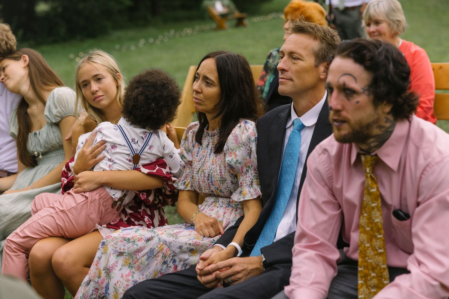 A group of people sitting together outdoors at a daytime gathering, wearing casual and formal attire.