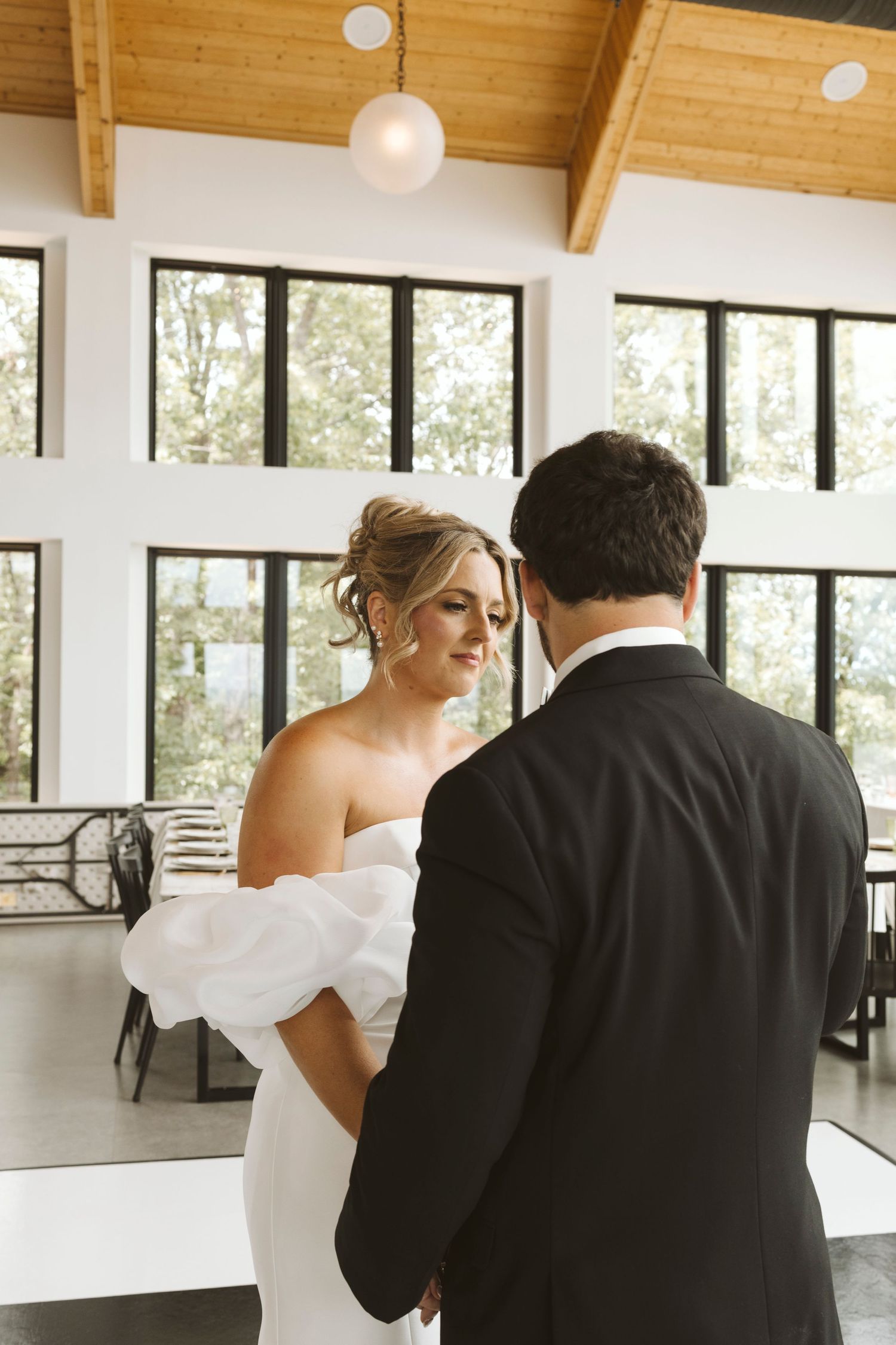 A bride and groom share an intimate moment in a modern venue with floor-to-ceiling windows and exposed wooden beams.