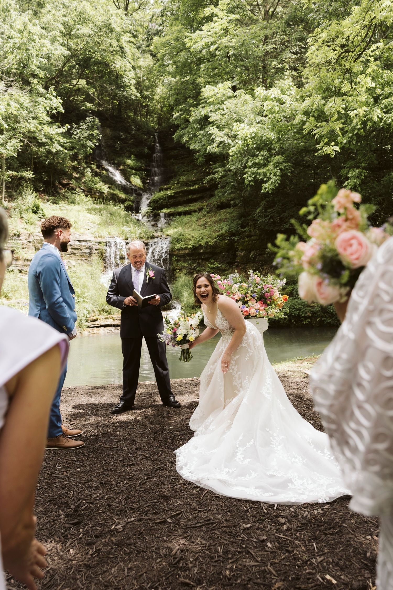 Bride in a flowing white dress stands with wedding party in a scenic wooded outdoor setting.