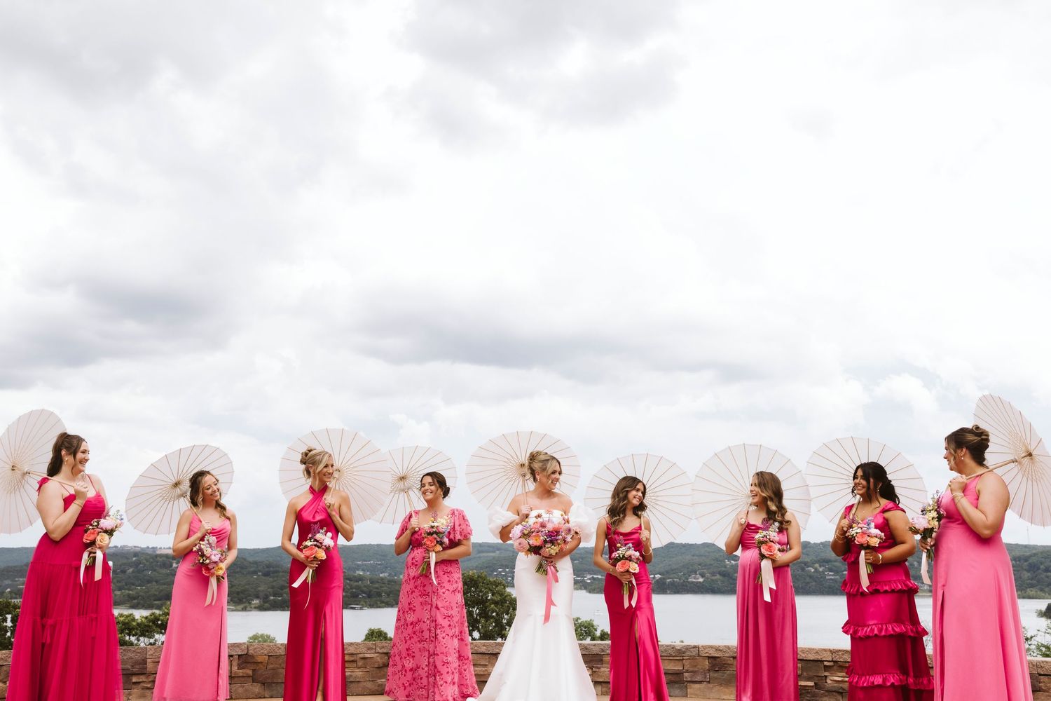 Group of bridesmaids in coral dresses lined up with bouquets for a creative panoramic wedding photo at outdoor venue.
