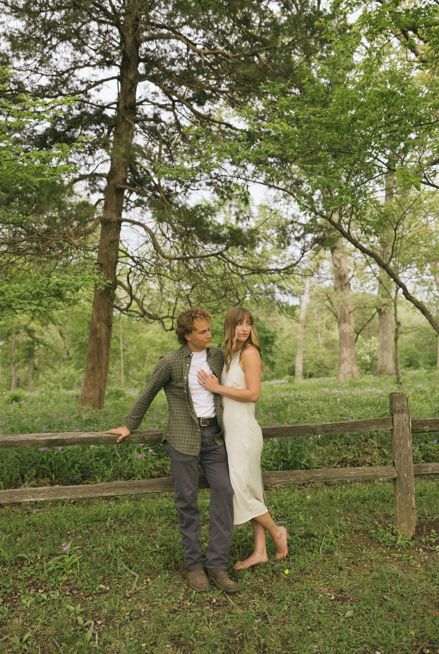 A romantic couple embraces by a rustic wooden fence in a lush green forest setting.