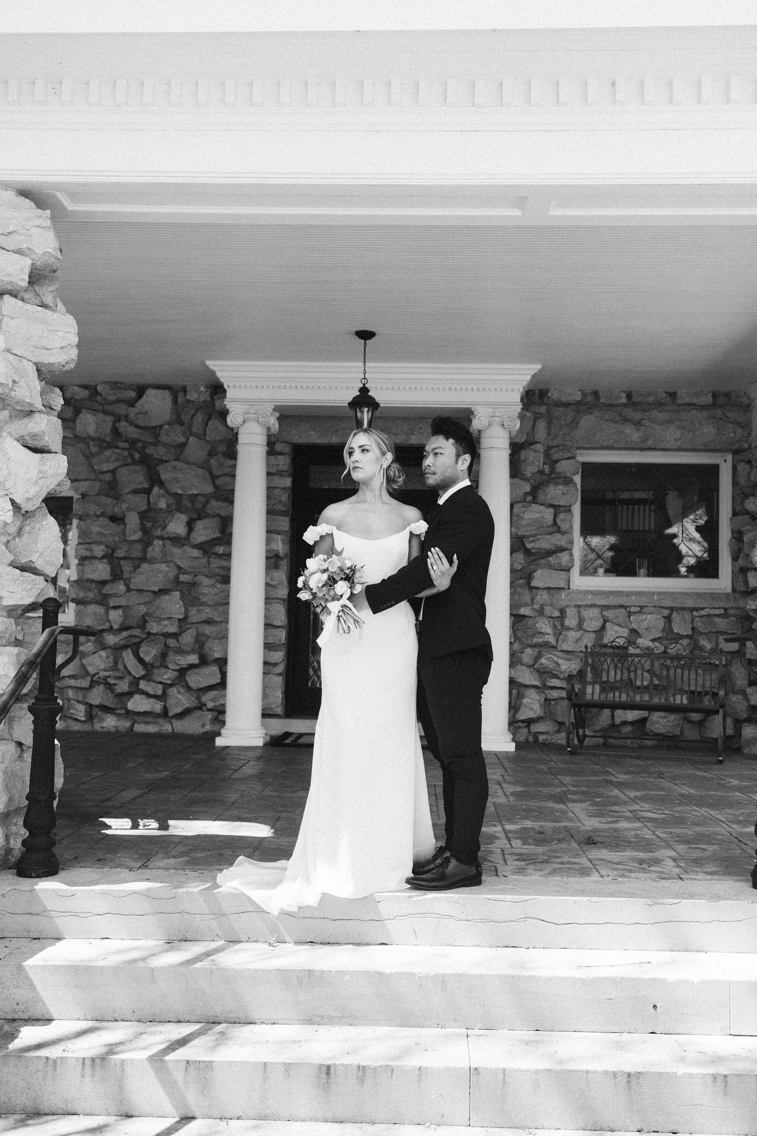 A bride and groom stand together on stone steps under white columns of a rustic stone building in black and white.