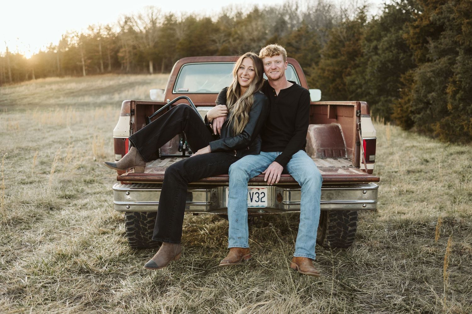 A couple sitting together in the bed of a vintage red pickup truck in a grassy field.