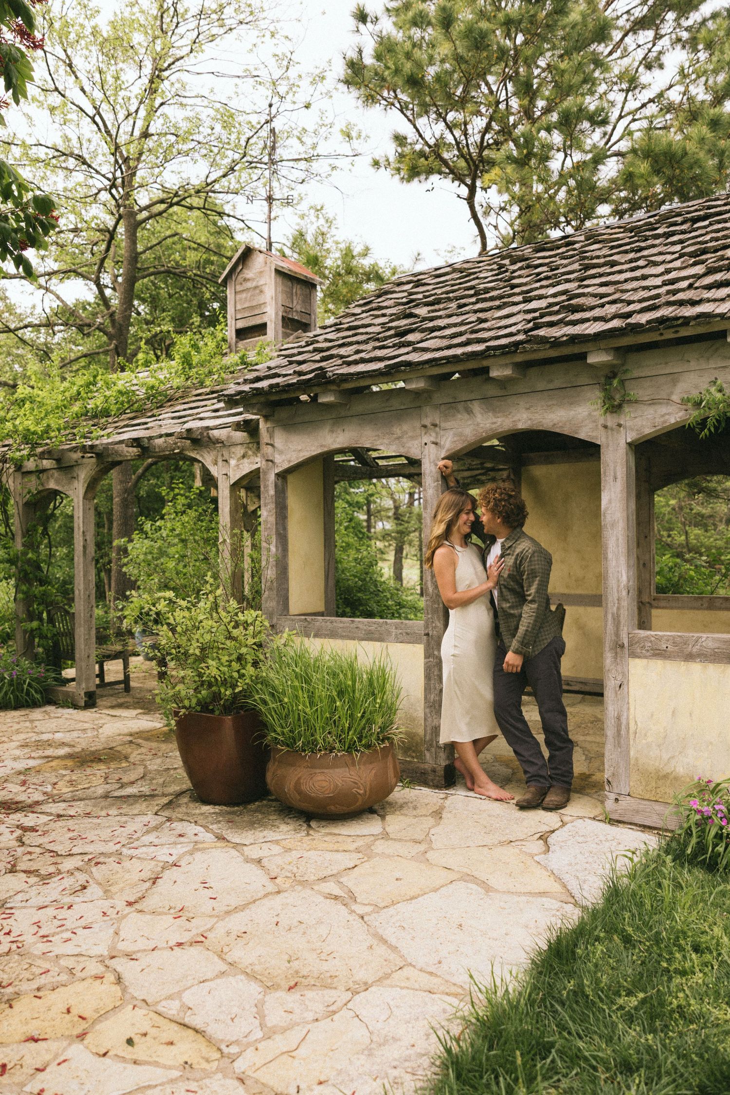 A romantic moment shared under a rustic pergola with stone pavers and potted plants.