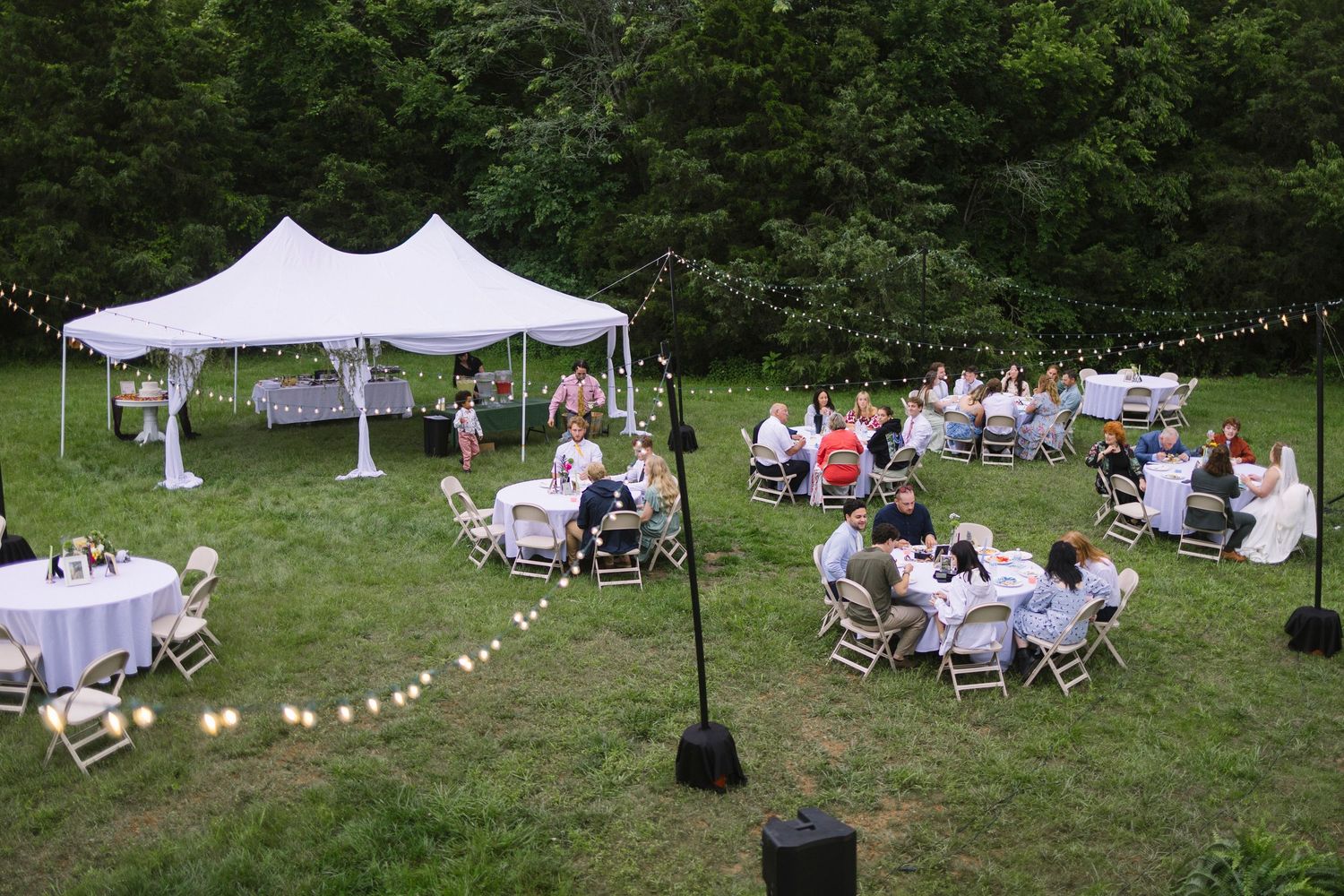 An outdoor wedding reception setup with white tents and string lights where guests are seated at round tables on a grassy lawn.
