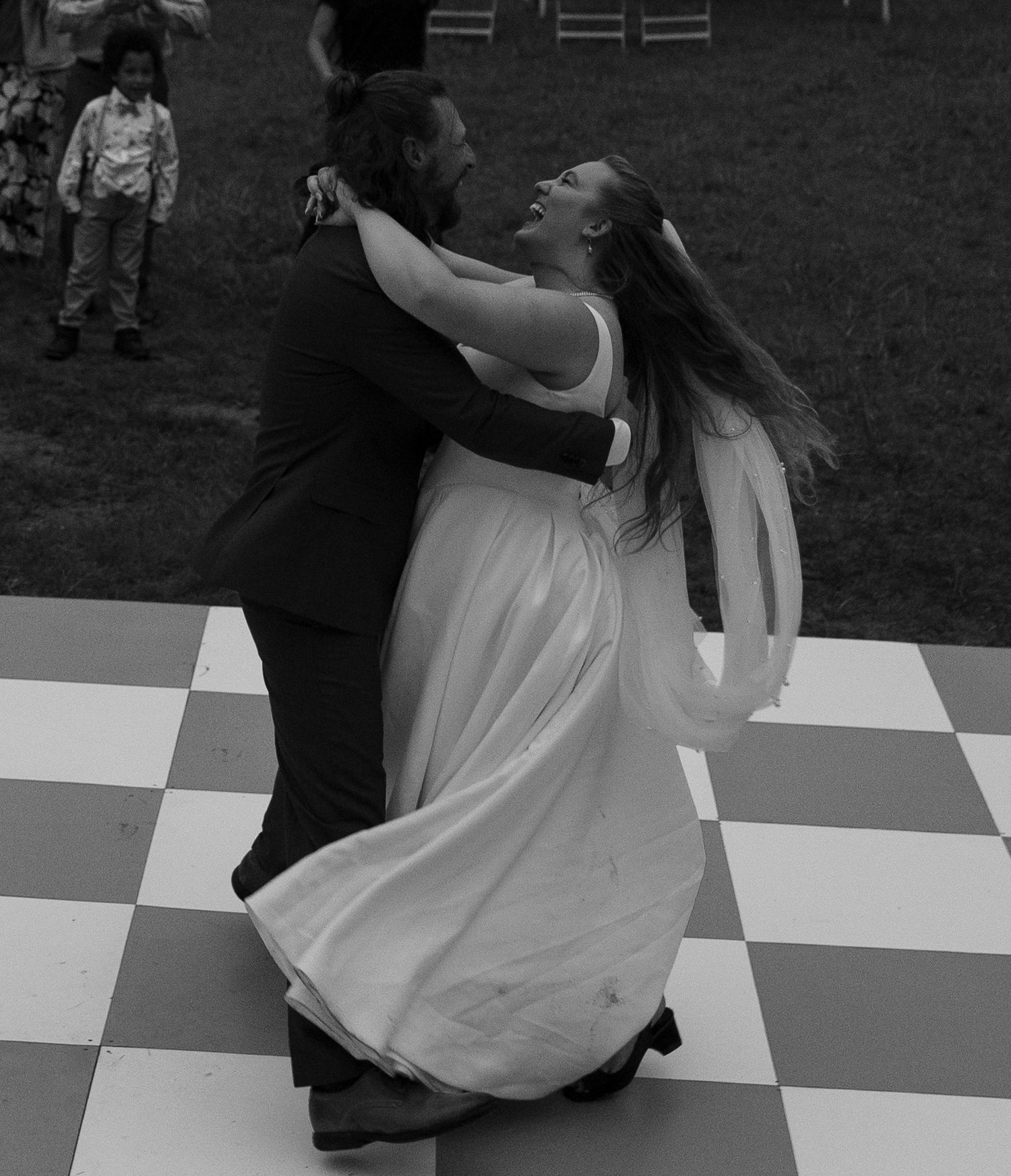 Two people dancing together on a checkered outdoor dance floor, with the bride in a flowing white gown and long veil.
