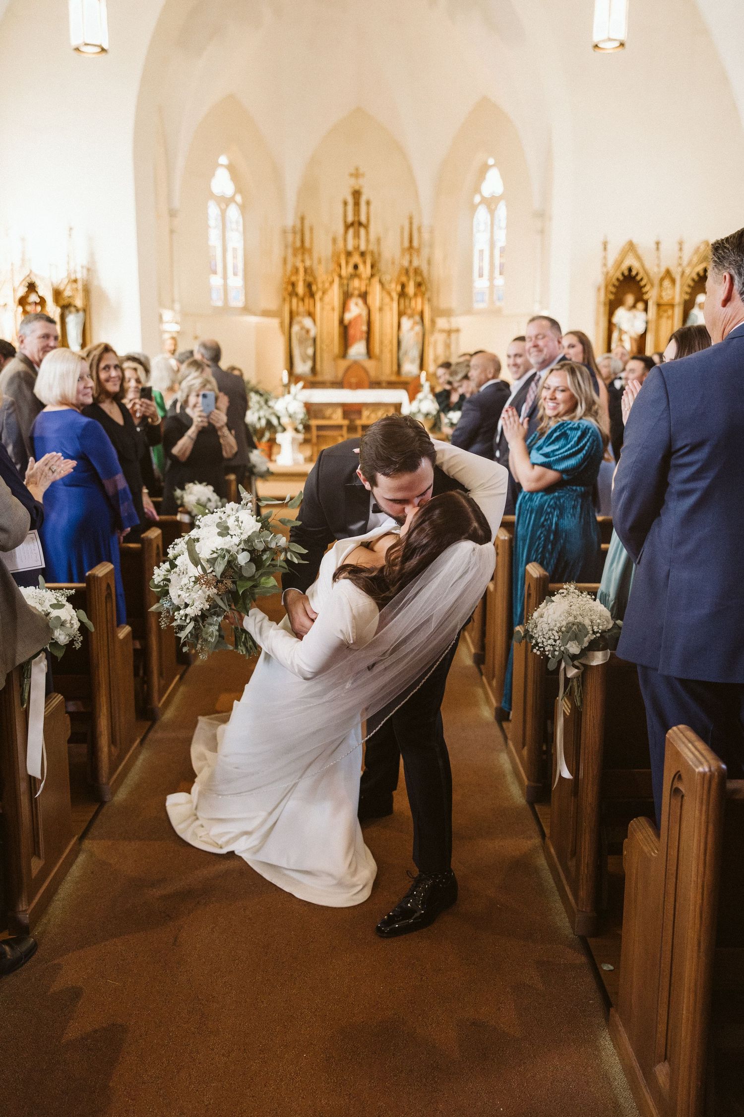 A newlywed couple shares their first kiss in the church aisle surrounded by wedding guests.