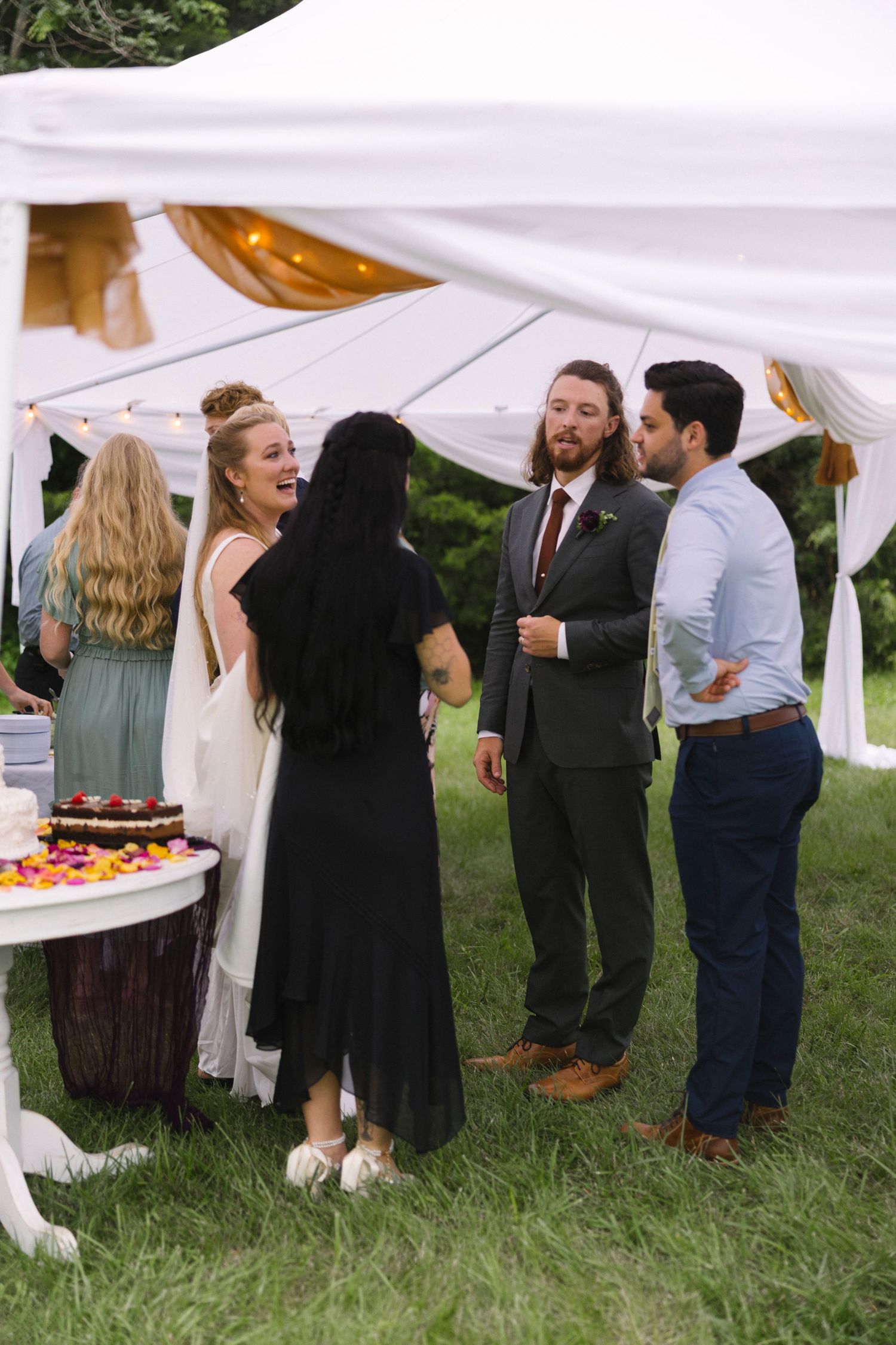 Groups of people mingling and socializing at an outdoor wedding reception under white tents on a grassy lawn.