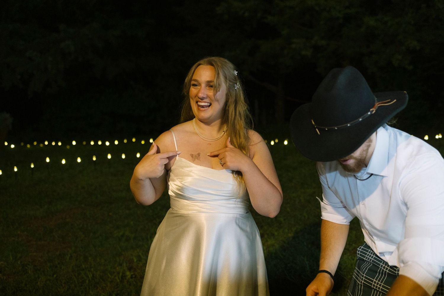 A bride and groom enjoy candid moments during a nighttime wedding reception under twinkling string lights.