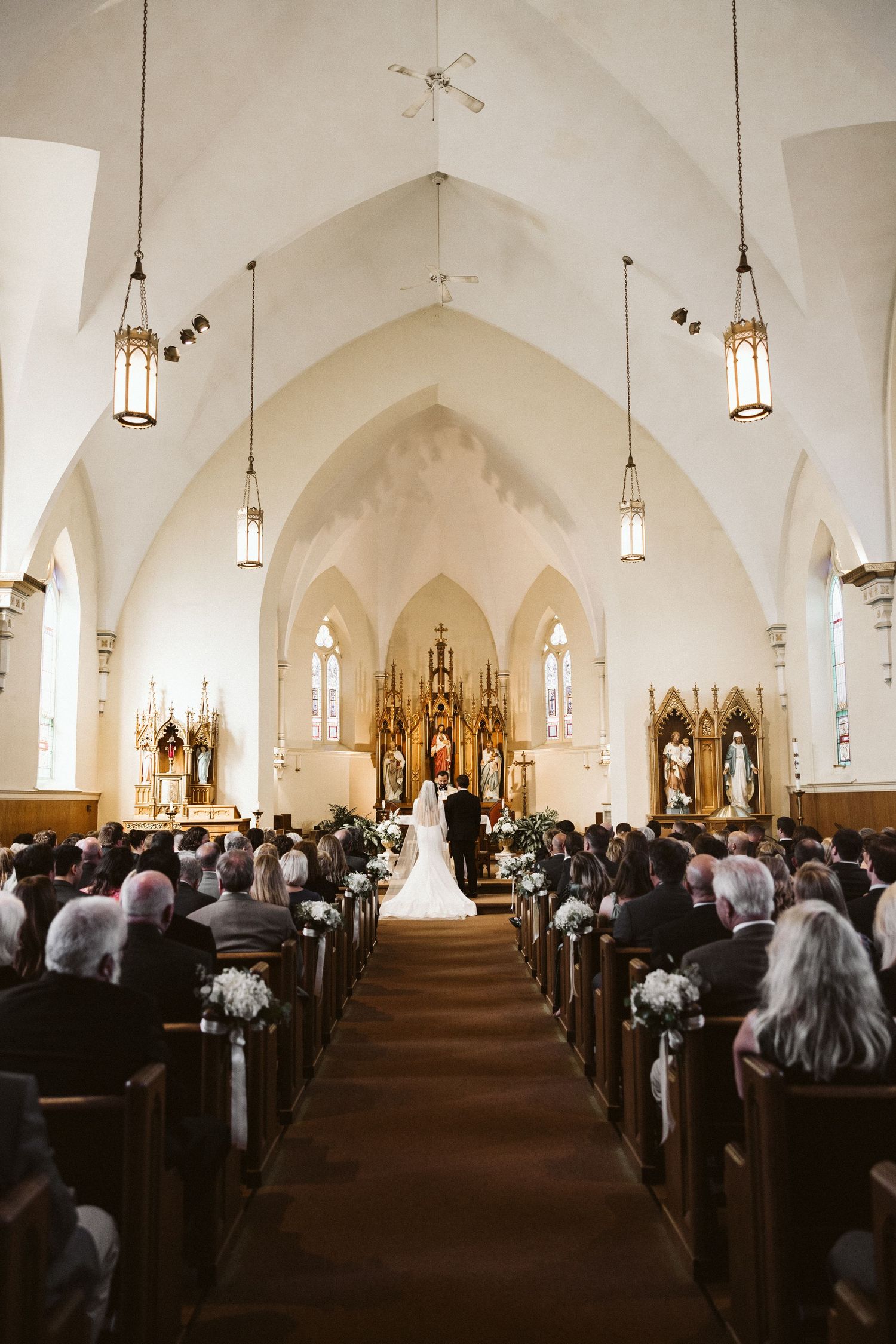 A white church interior with gothic arches and hanging lanterns during a wedding ceremony.
