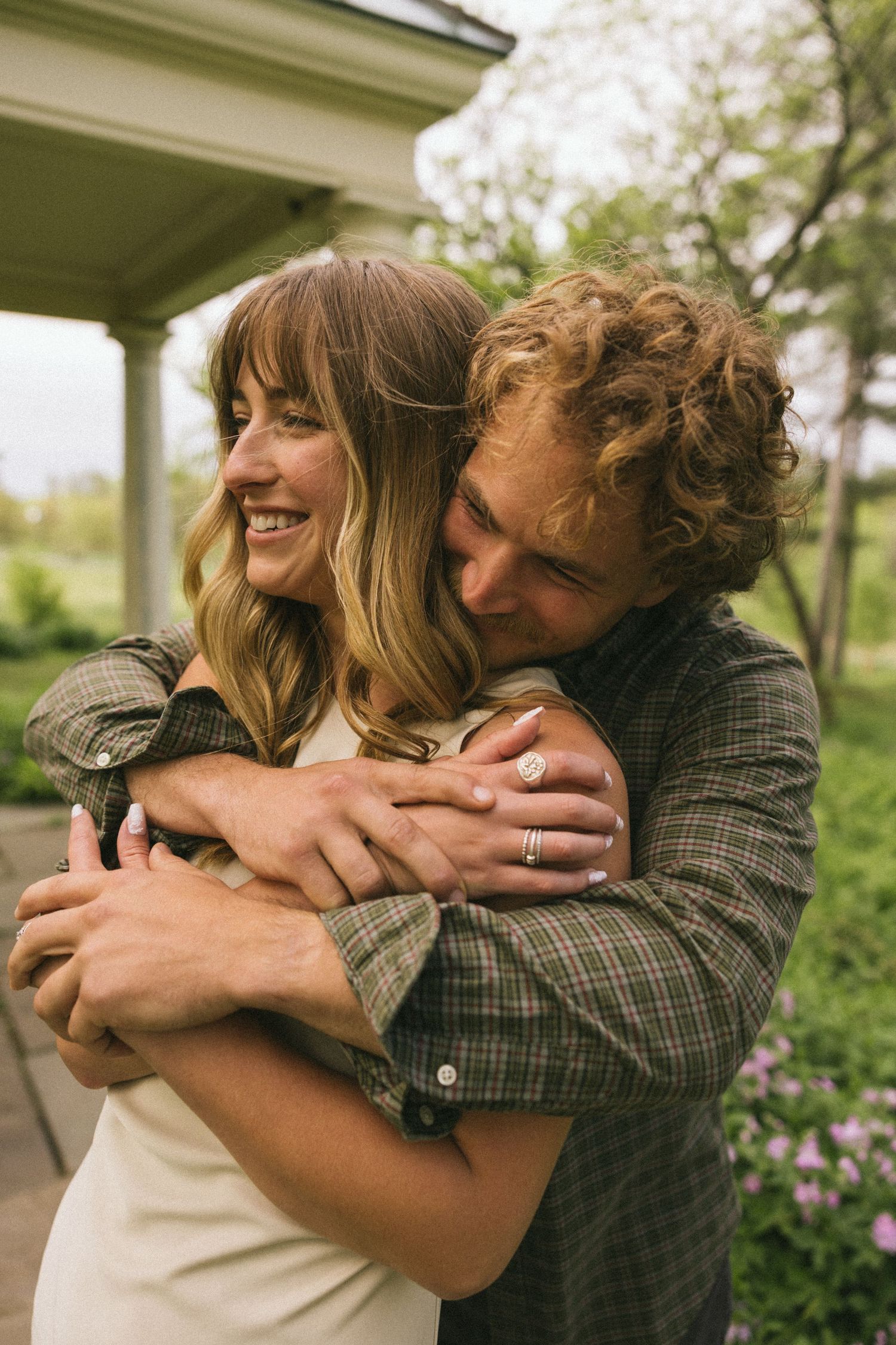 Two people share joyful embraces in a garden setting with flowering plants visible.