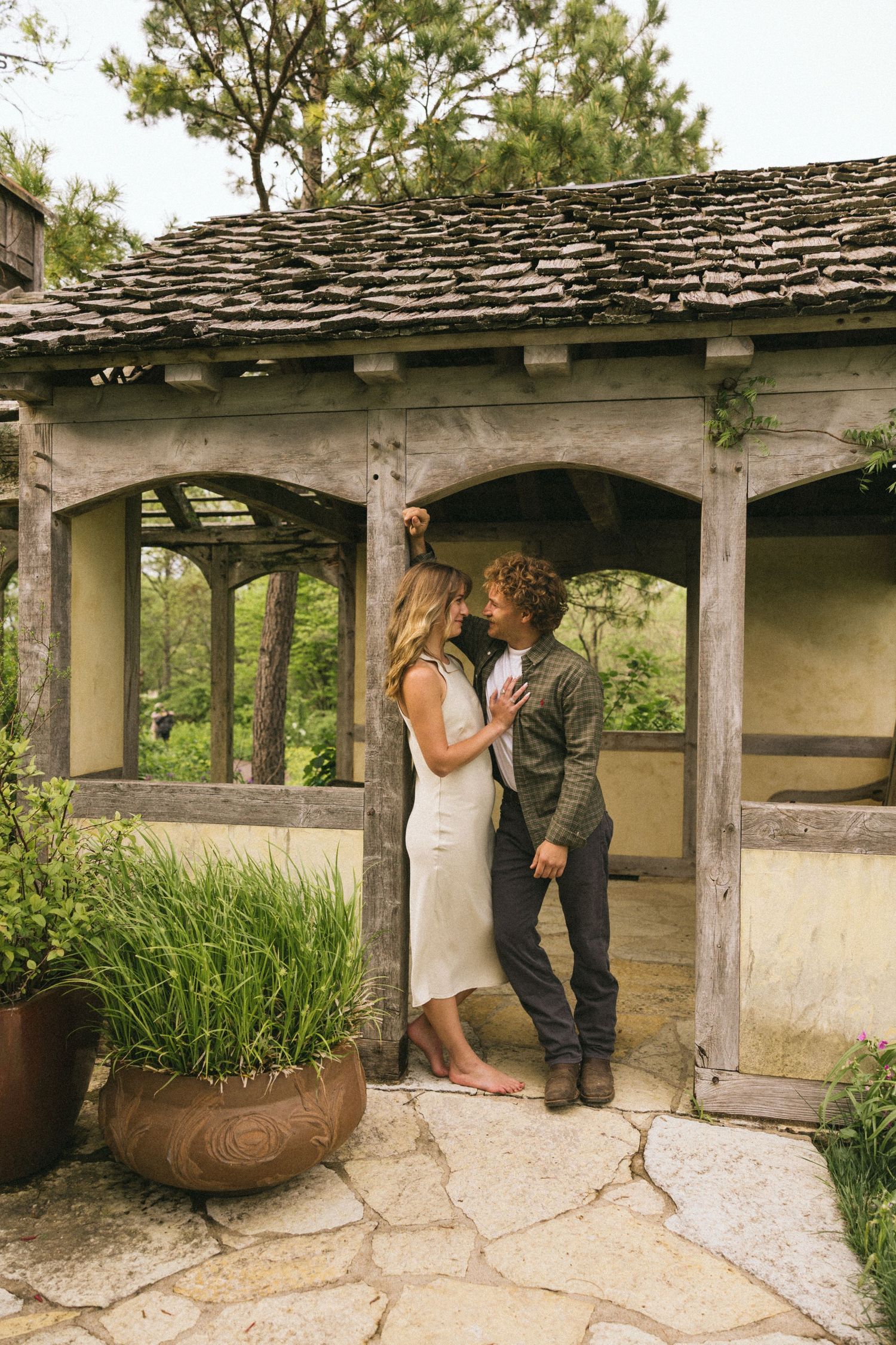 A couple shares an intimate moment under a rustic stone gazebo with a weathered shingle roof.