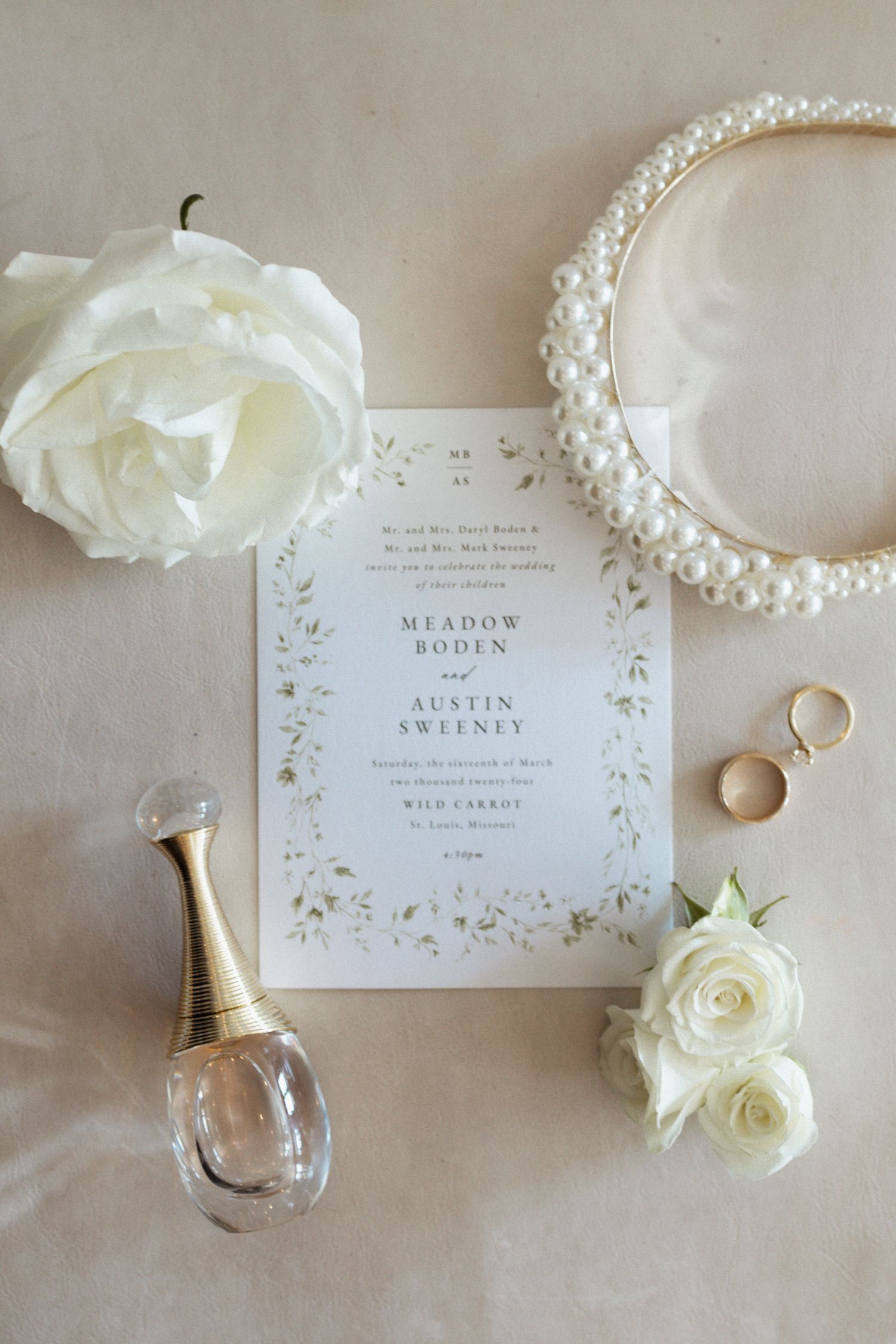 Wedding invitation and bridal accessories including pearl headband, white roses, perfume bottle and gold rings on beige surface.