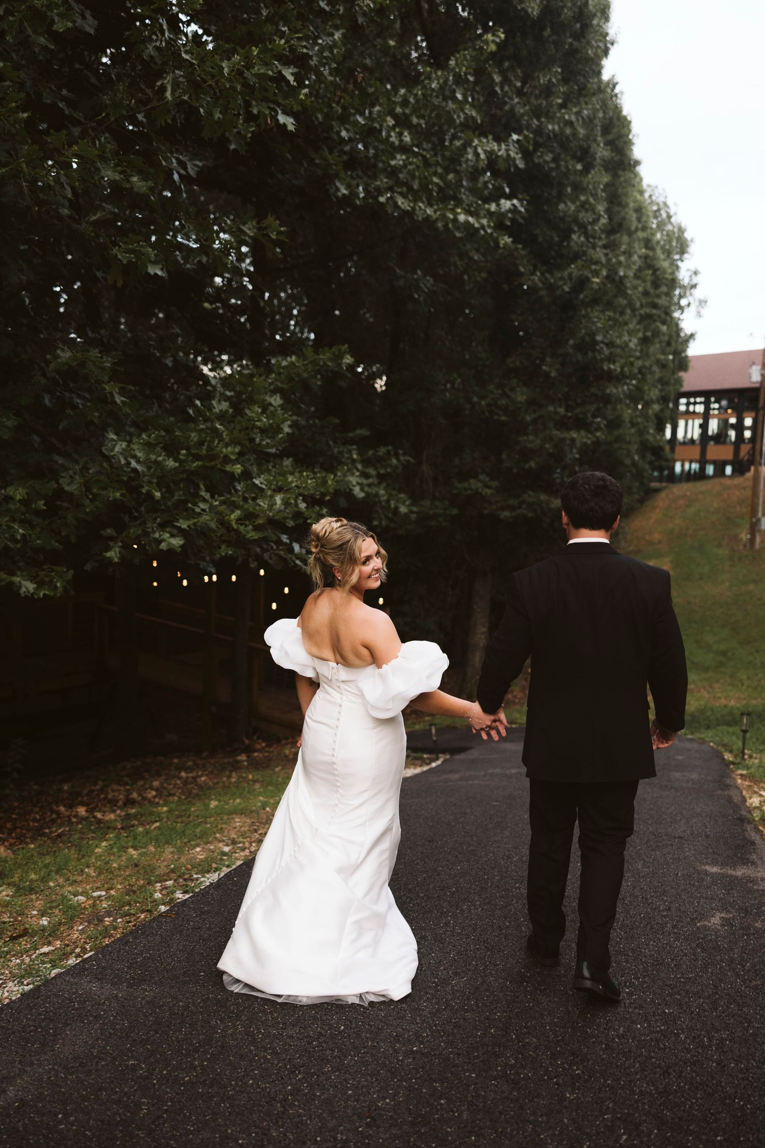 A couple walking together down a tree-lined path with the bride holding up the skirt of her white wedding dress.