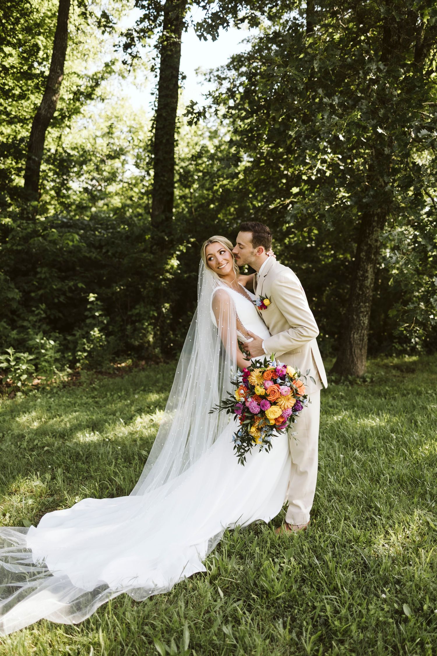 Couple shares intimate moment in wooded setting with colorful cascading bouquet.