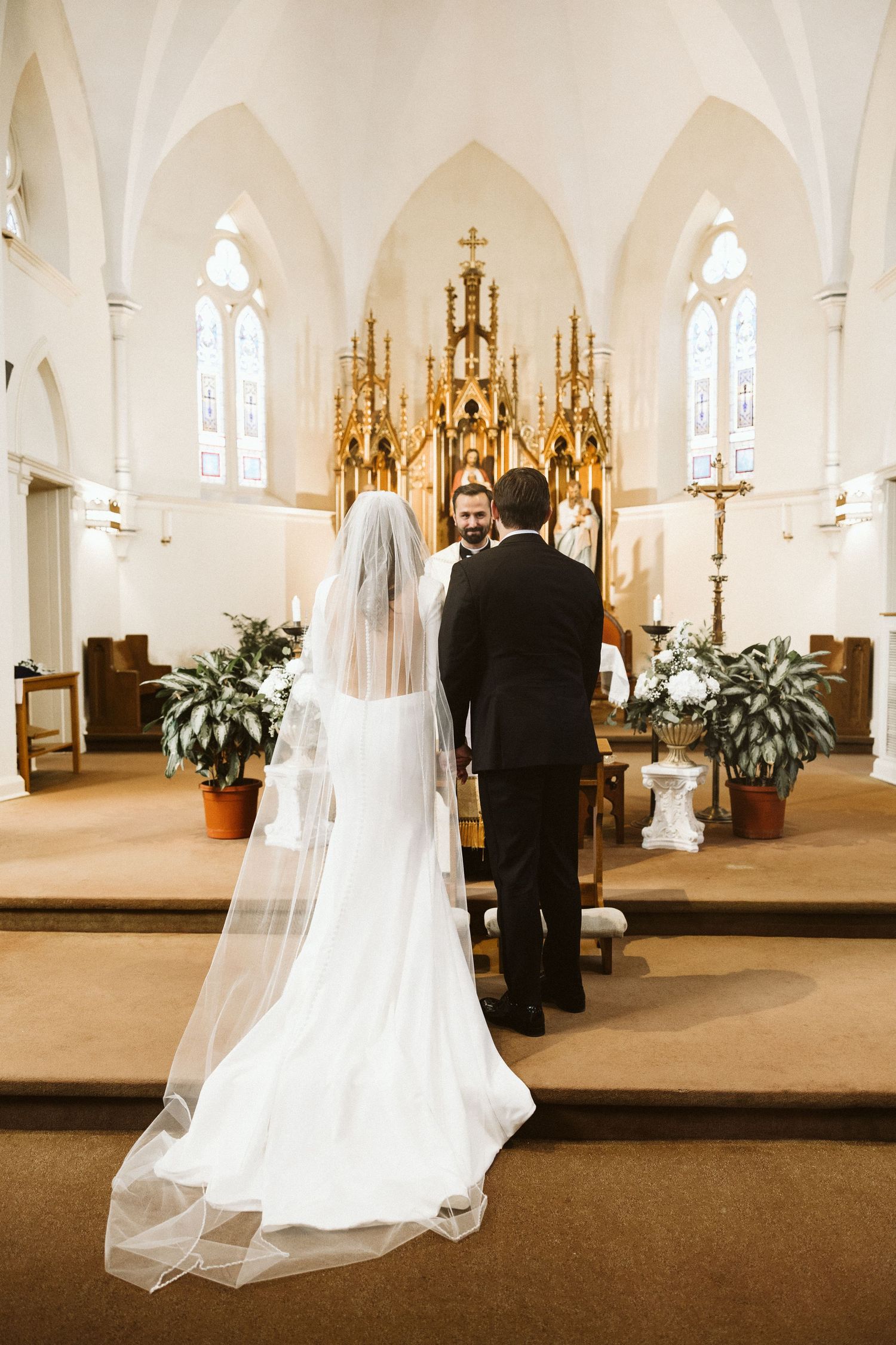 Couple walking down church aisle with Gothic altar and stained glass windows in background.