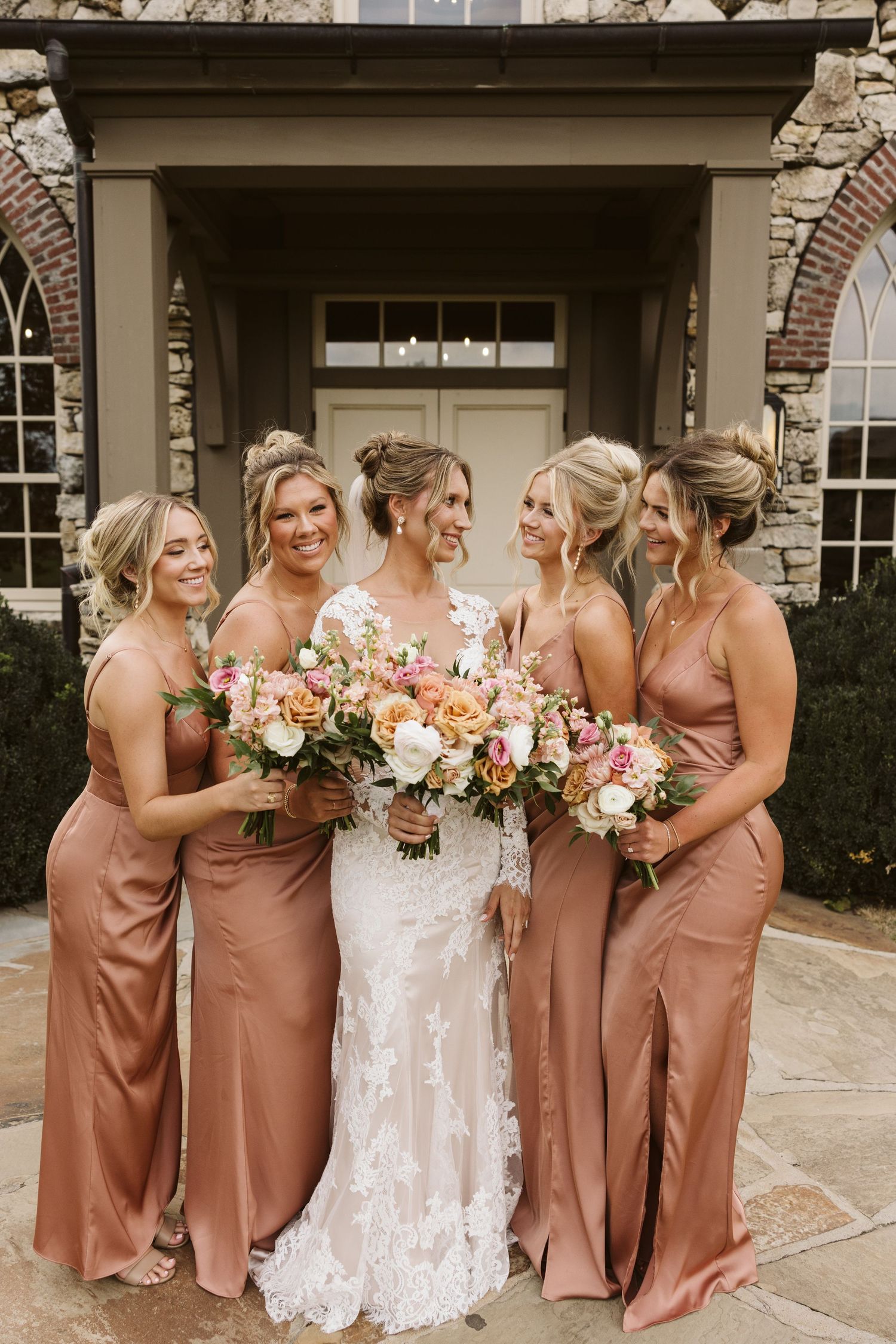 Wedding party members in silk rose-colored dresses gather for photos outside a stone church.