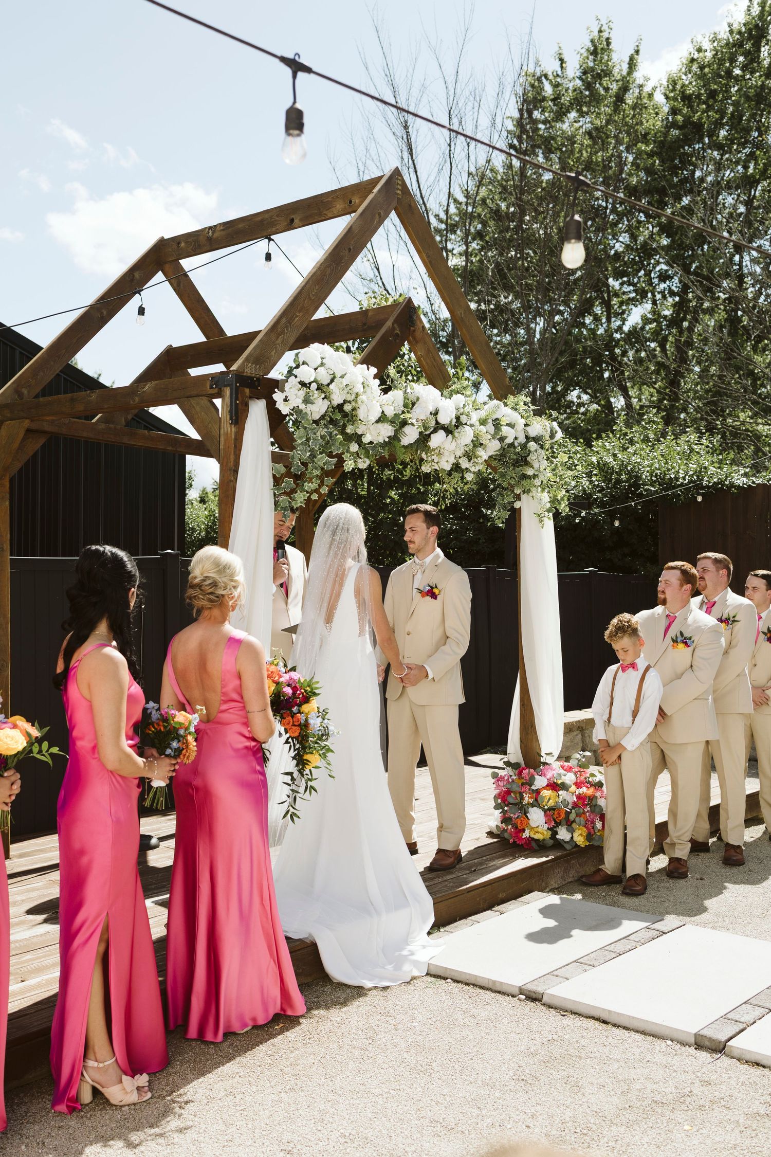 Wedding ceremony setup with wooden pergola decorated in white flowers and string lights.