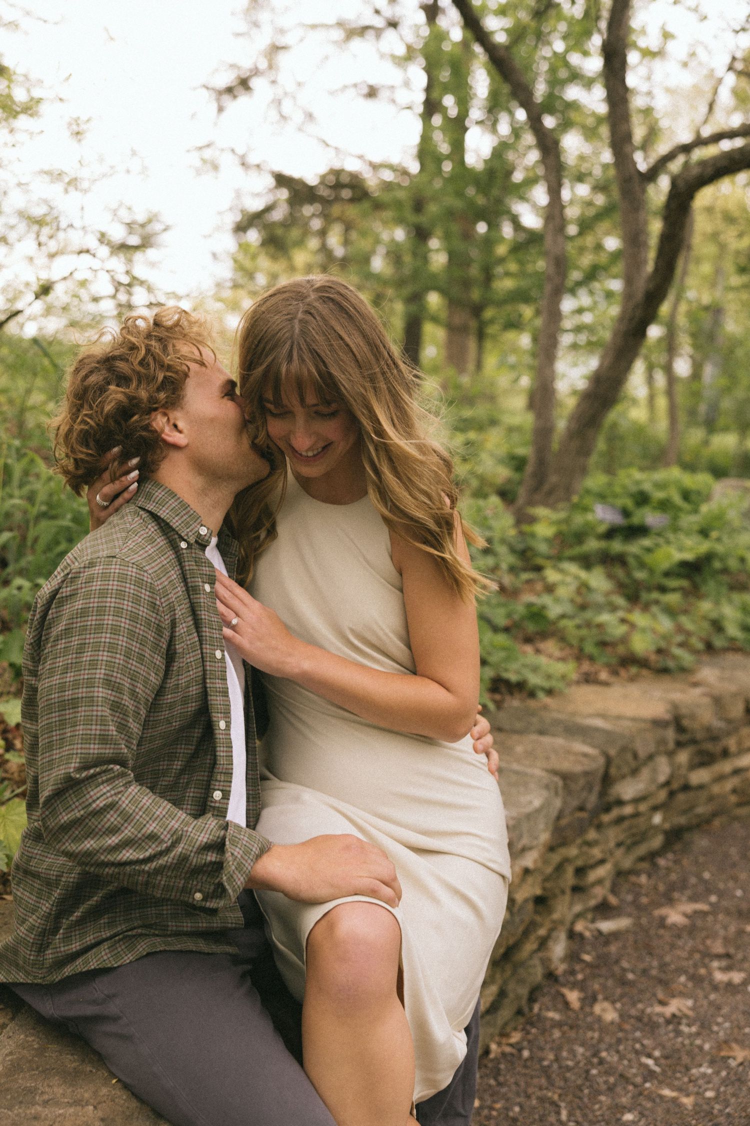 A tender moment between a couple sitting on stone wall in a wooded garden setting.