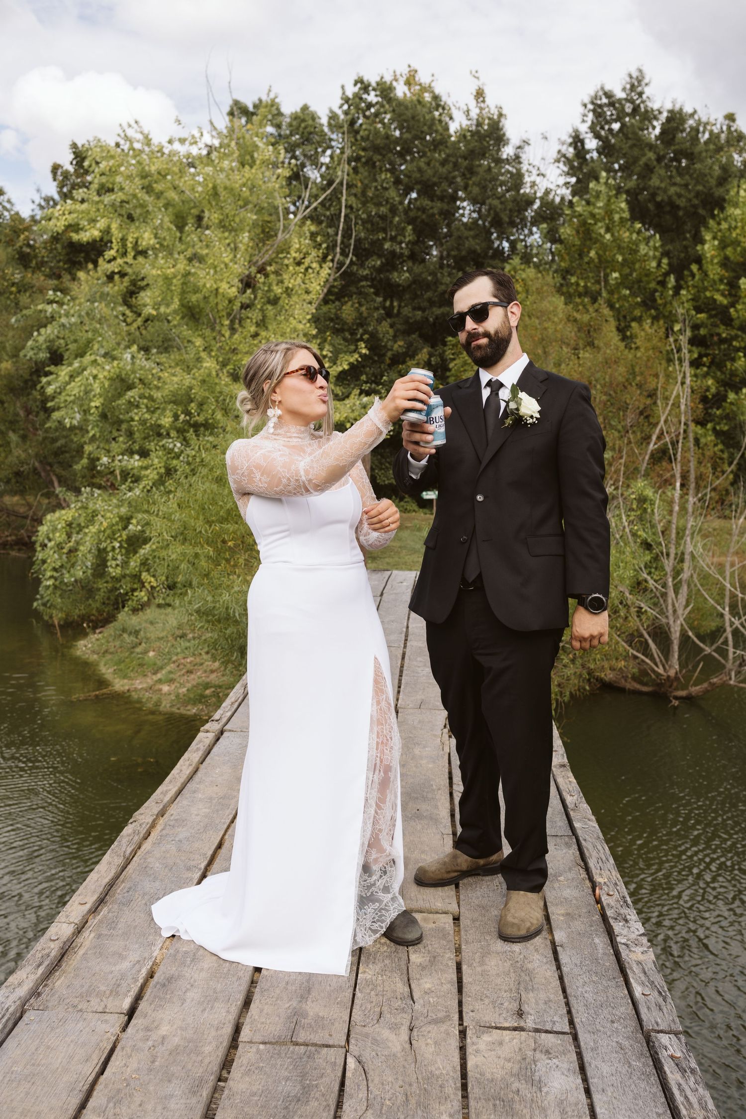 Wedding couple toasts with drinks while standing on a rustic wooden dock by a peaceful lake.