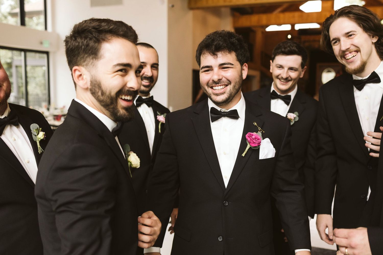 Groomsmen in black tuxedos and bow ties sharing a candid laughing moment at a formal wedding celebration.