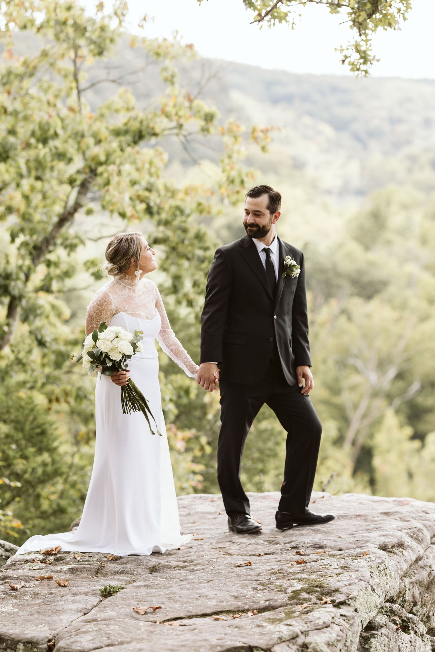 Romantic wedding couple poses together on a scenic rocky overlook surrounded by trees and mountains.
