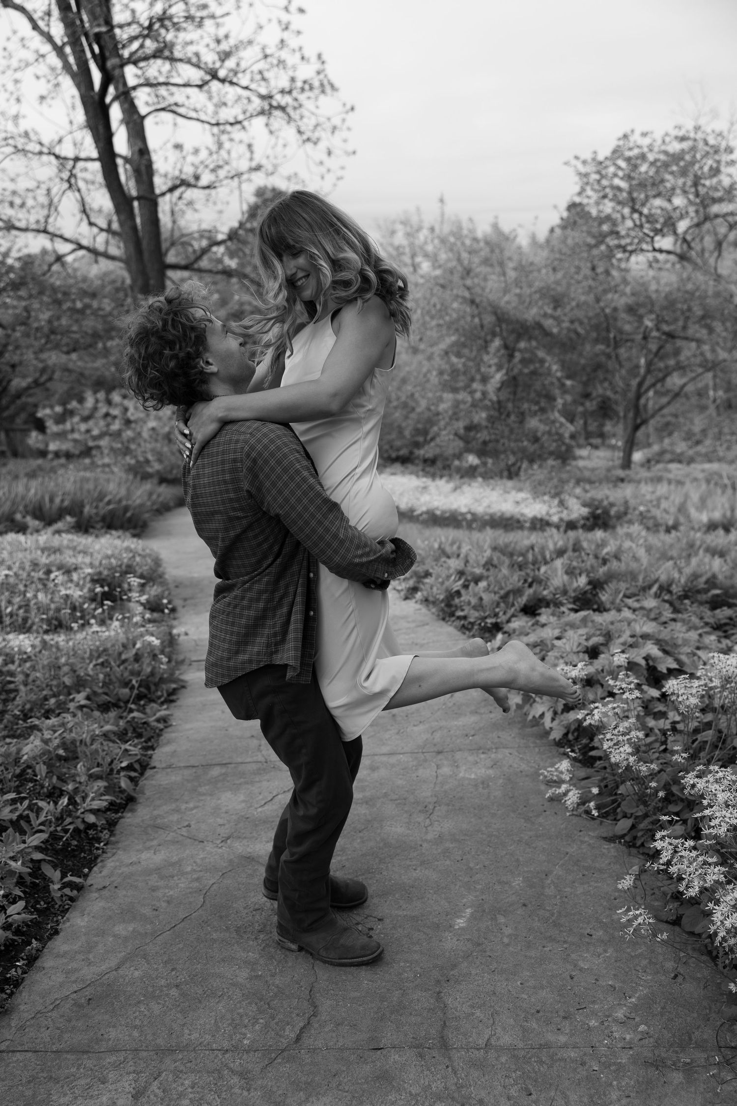 A joyful couple embracing on a stone pathway surrounded by gardens and trees.