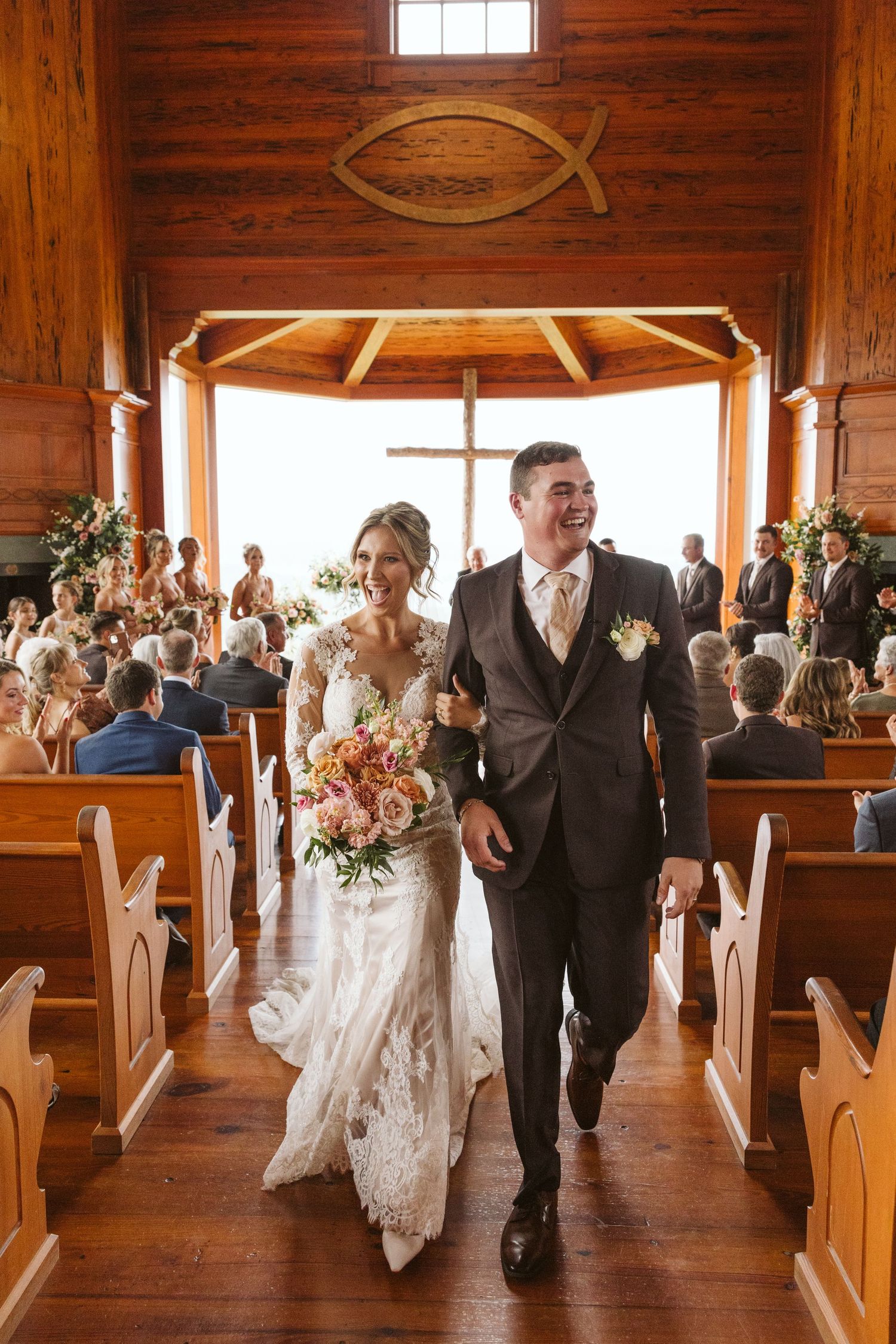 A newlywed couple walking down the aisle of a rustic wooden chapel with a Christian fish symbol overhead.