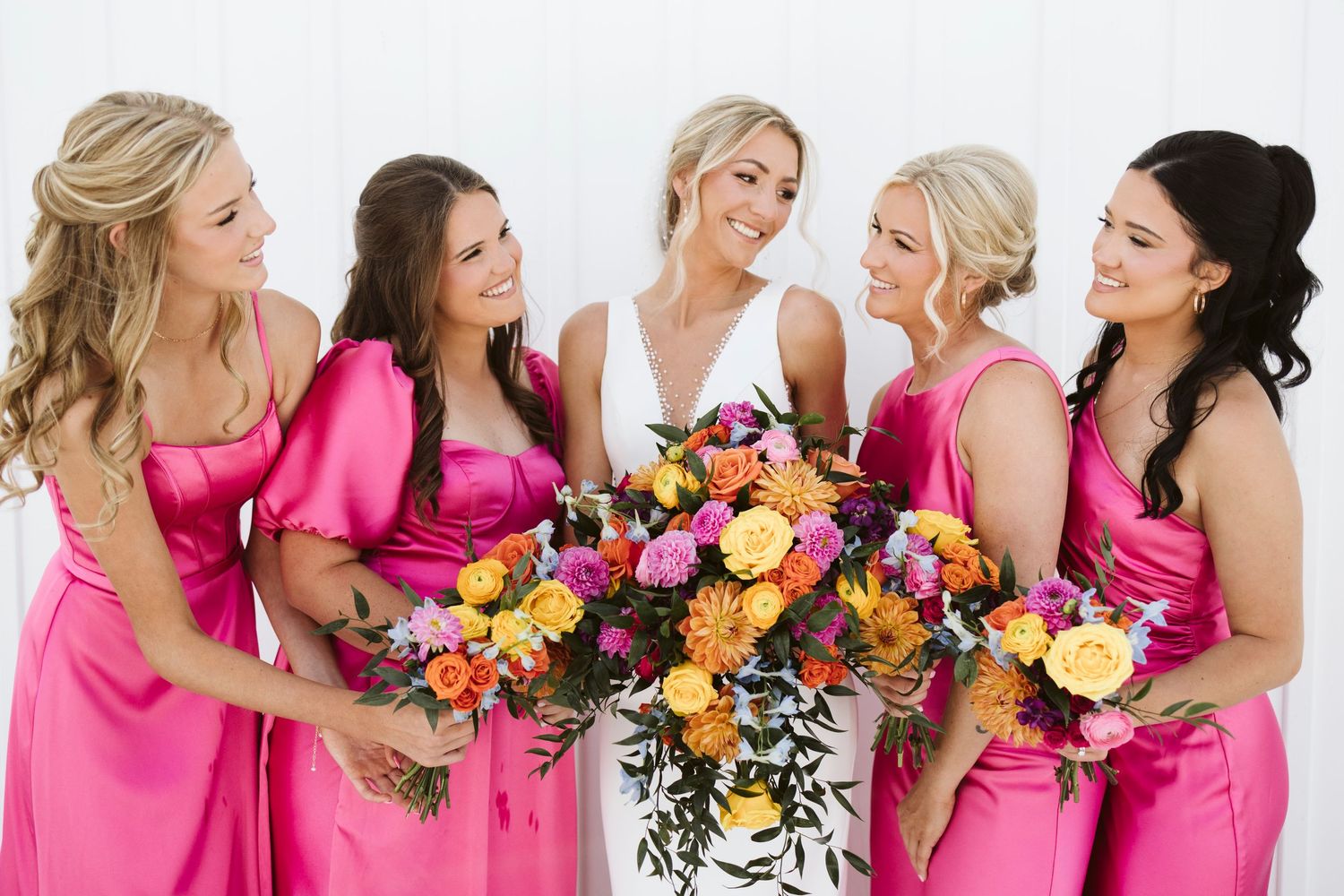 Bridal party in hot pink dresses share candid laughs while holding vibrant floral bouquets.