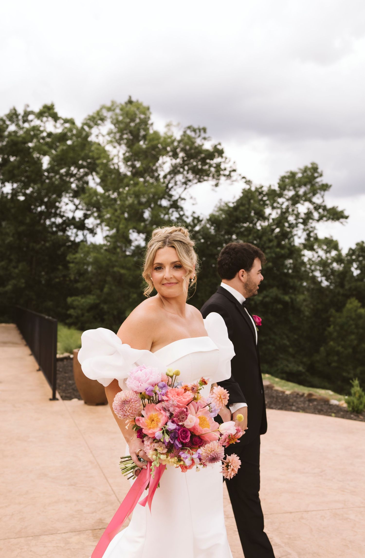 Two wedding attendees pose outdoors on a stone path with beautiful greenery and cloudy skies in the background.