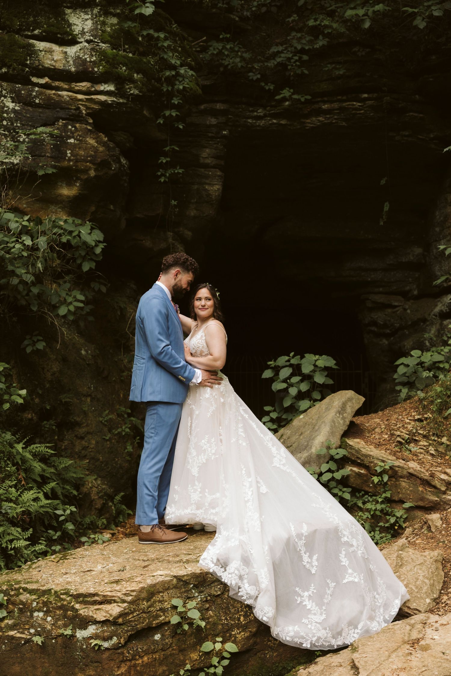 Couple in wedding attire poses together against dramatic rock formations in a cave-like setting.