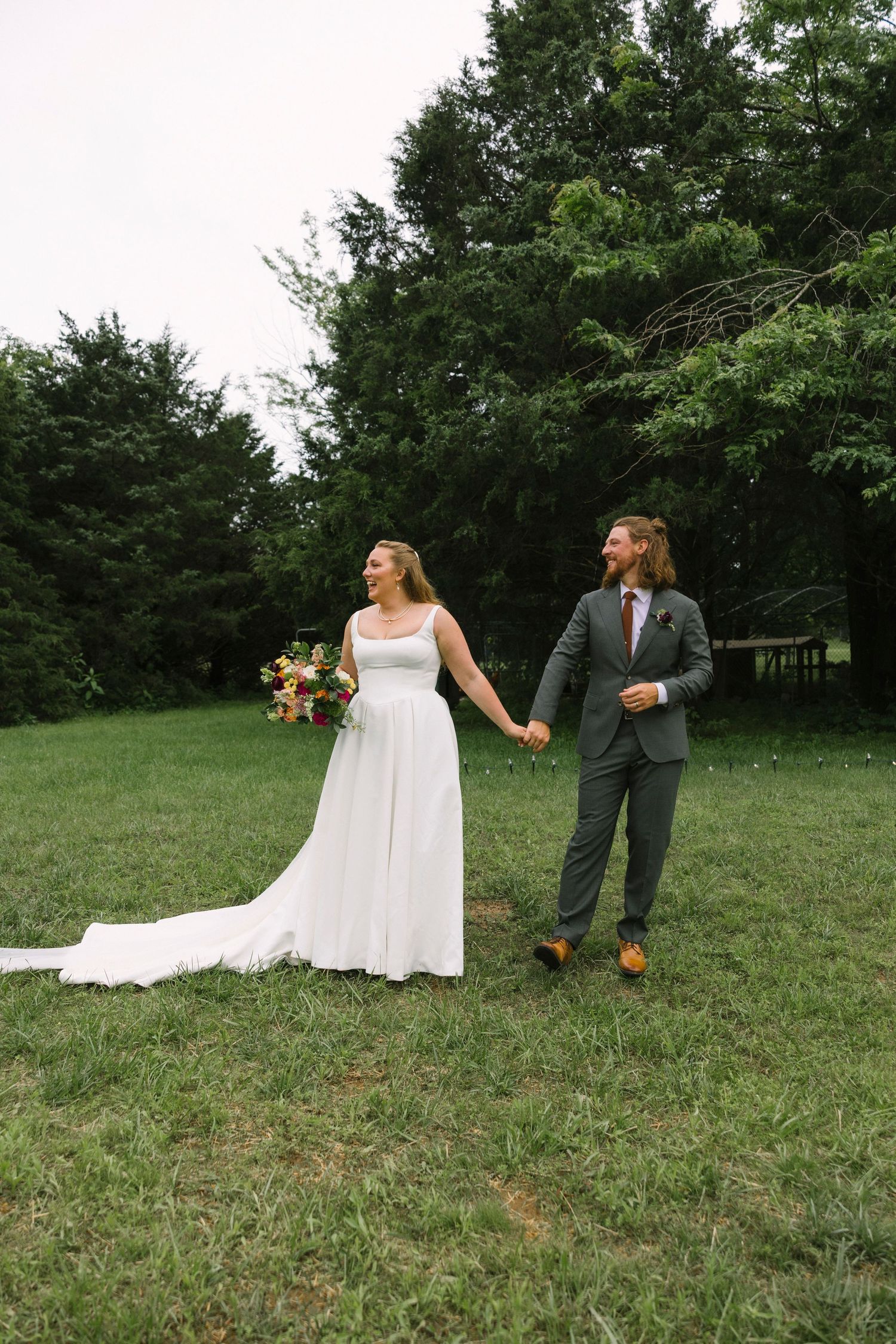 A couple in wedding attire walks hand-in-hand across a green lawn with trees in the background.