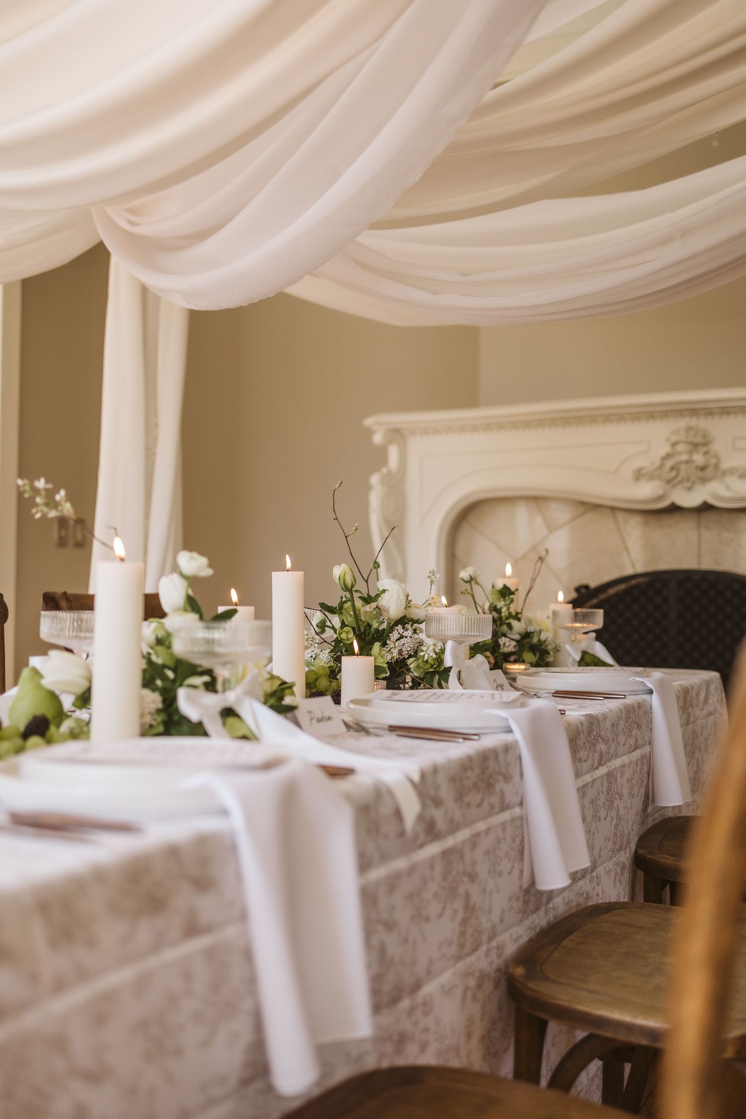 An elegant wedding table setting with white linens, draped fabric, candles and floral arrangements near an ornate fireplace.