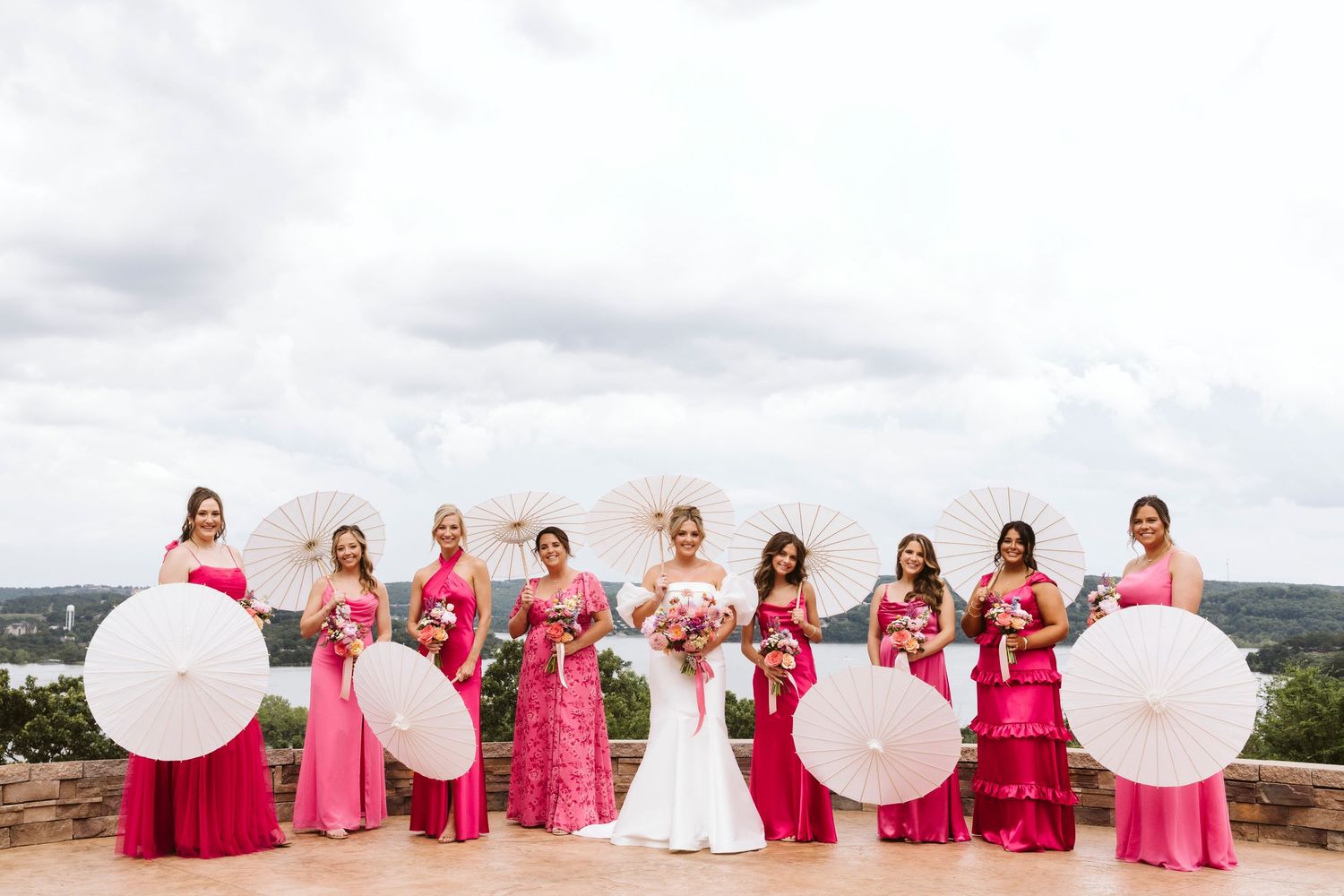 Wedding party in pink and red dresses pose with large white balloons on outdoor terrace overlooking scenic landscape.