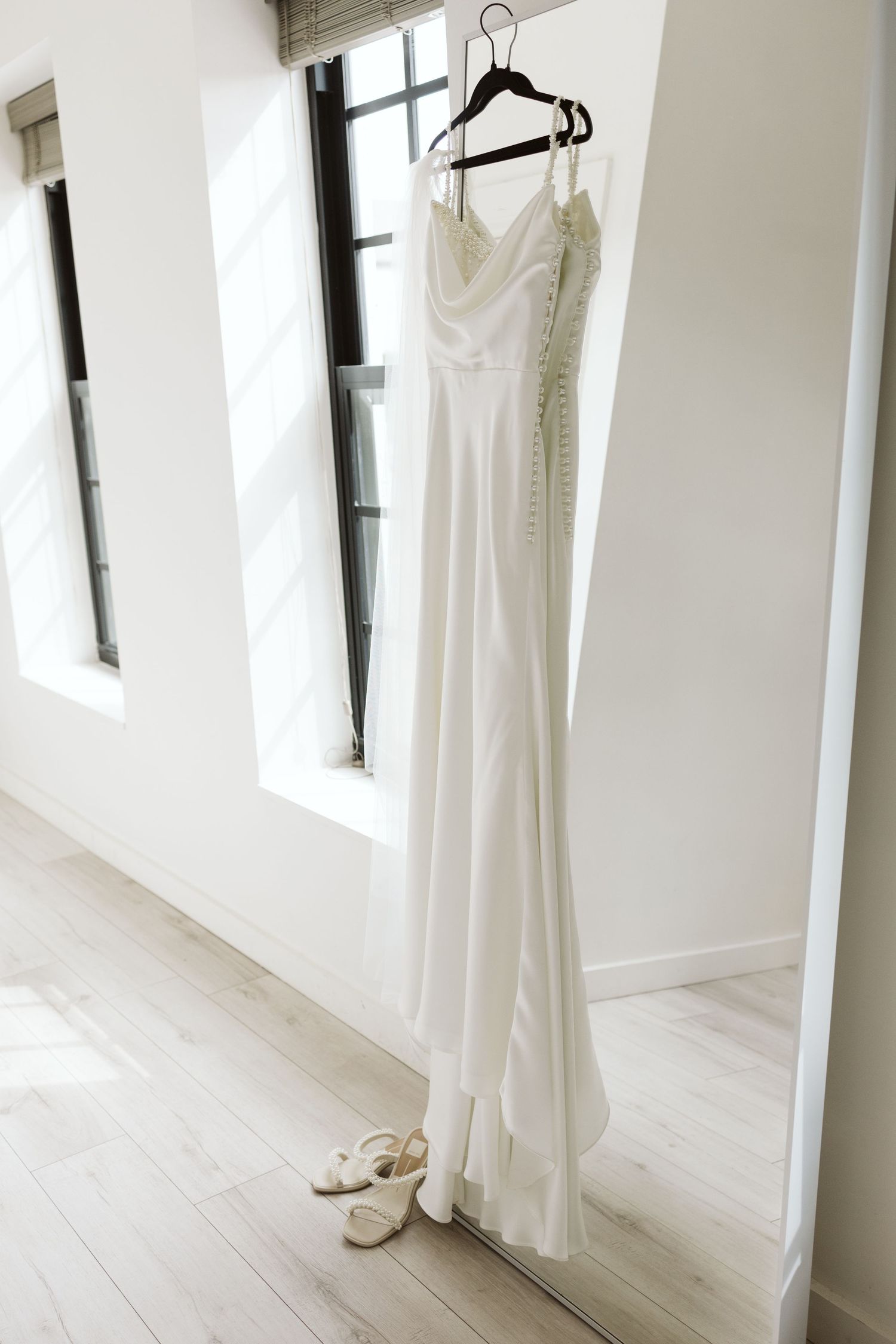 White wedding dress hanging on display with matching bridal shoes on a light wooden floor in a bright room.