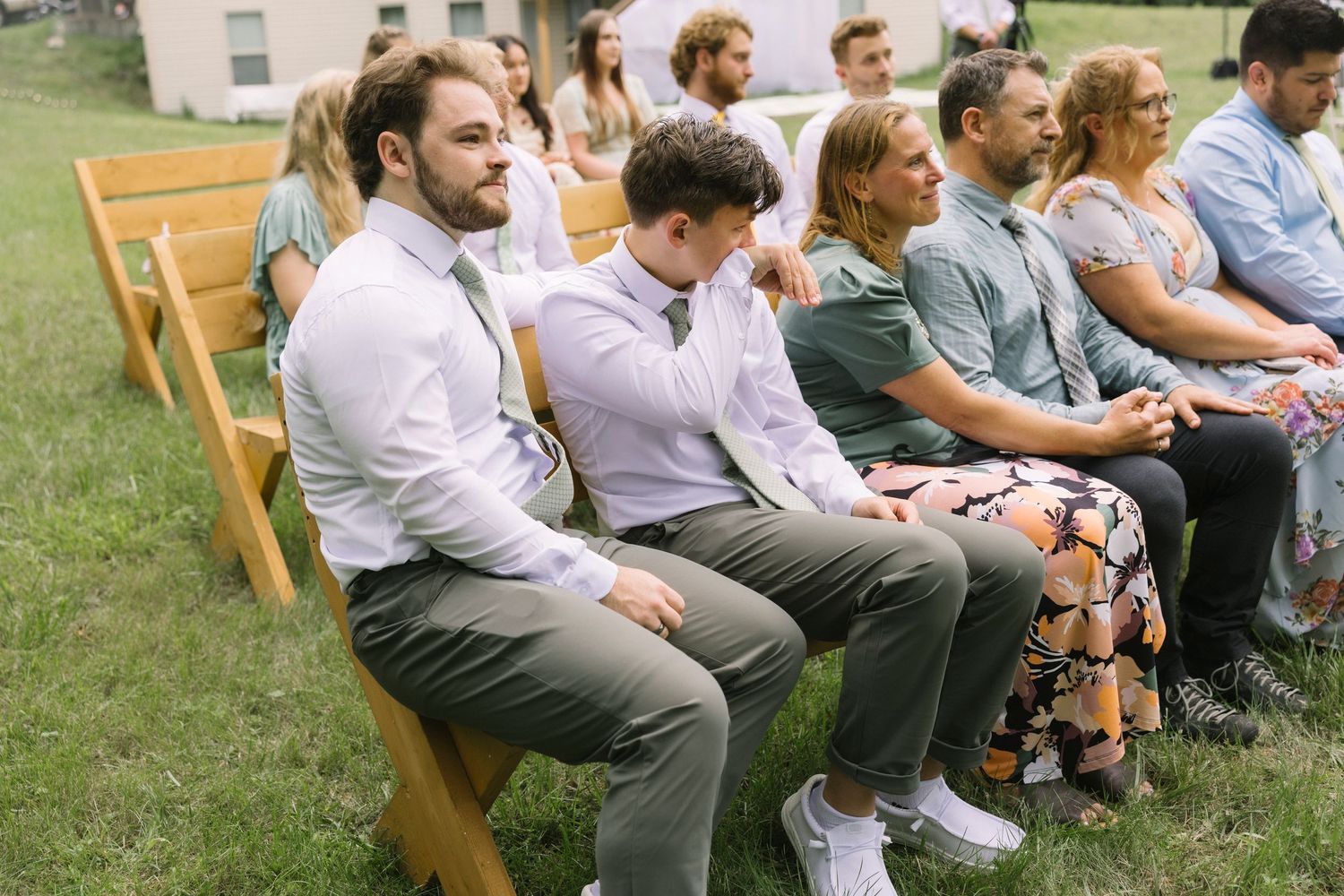 Guests seated on wooden folding chairs in rows on a grassy lawn at an outdoor ceremony or gathering.