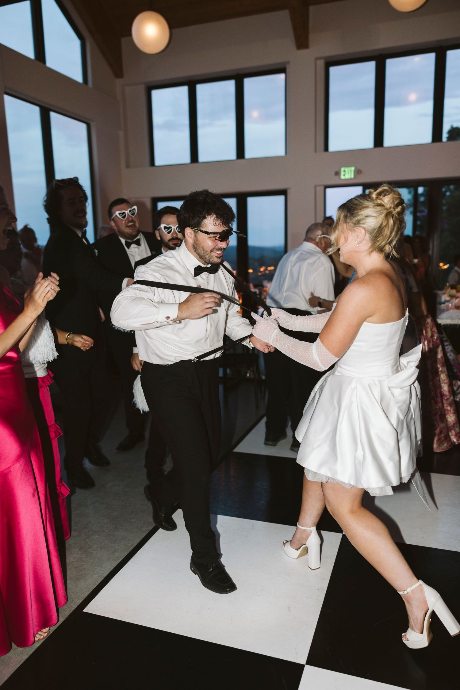 Wedding guests dance together on a black and white checkered dance floor at an indoor reception with large windows.