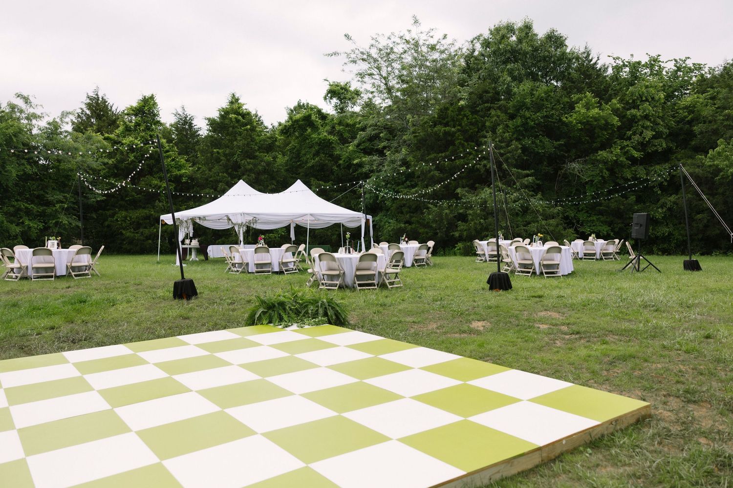 Outdoor wedding reception setup with white tent, tables, and large green and white checkered dance floor in grassy field.
