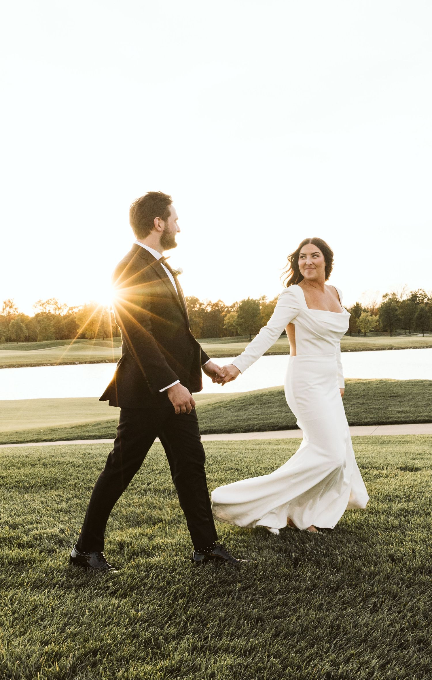 Couple in wedding attire playfully running through a grassy field at sunset.