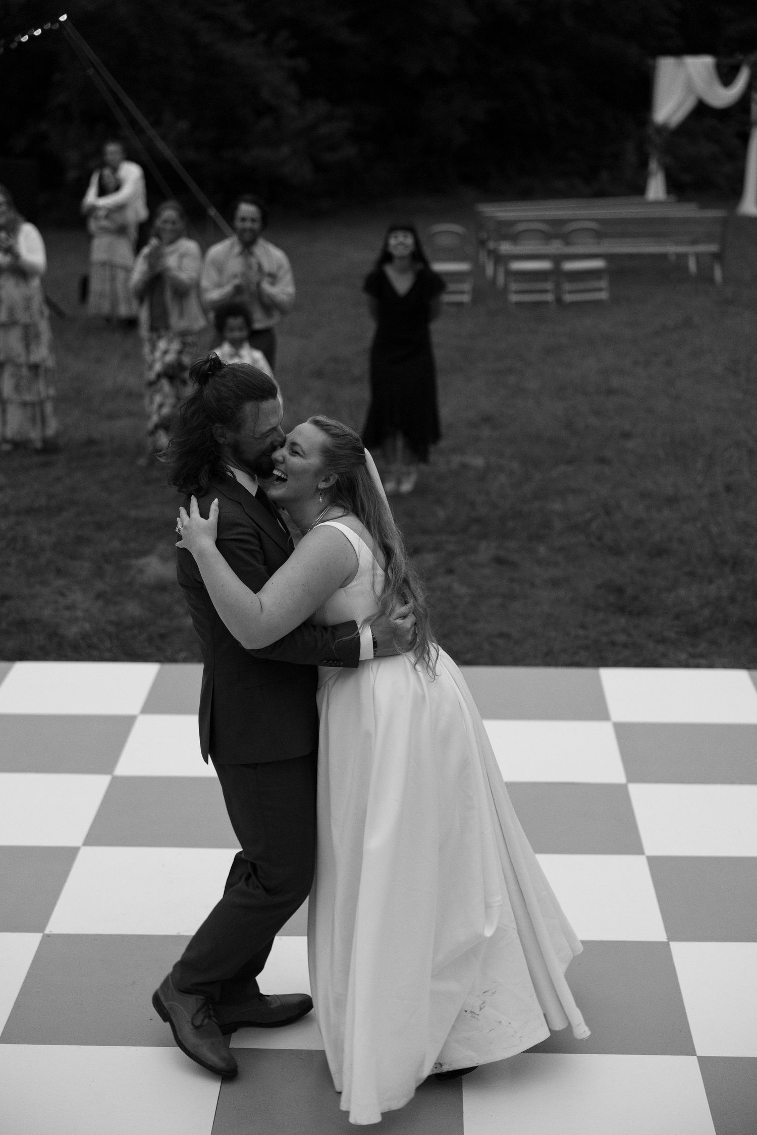 A couple dances together on a black and white checkered dance floor at an outdoor wedding.