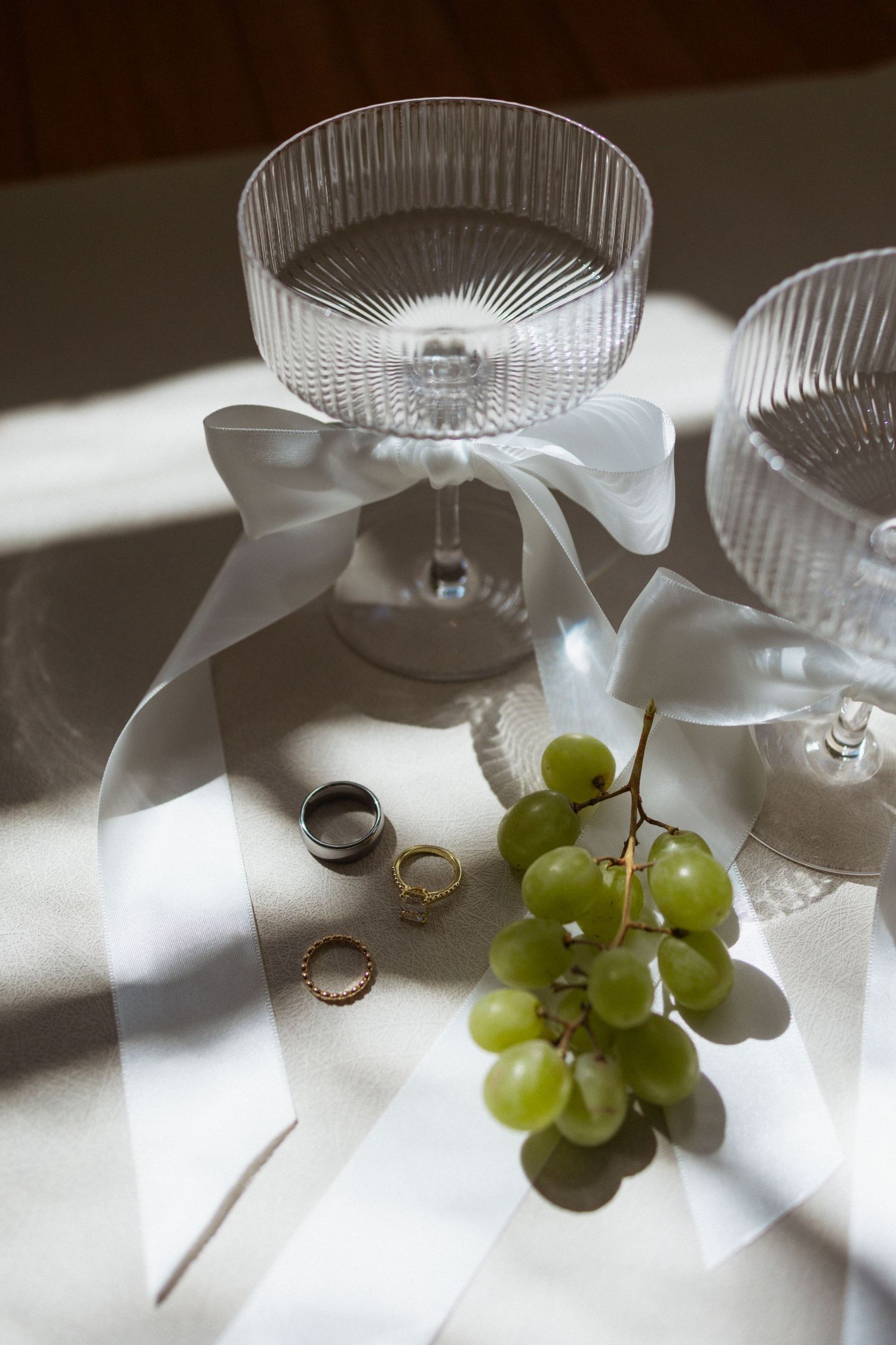 Elegant crystal champagne coupes with white ribbons, wedding rings and fresh green grapes in dramatic natural lighting.