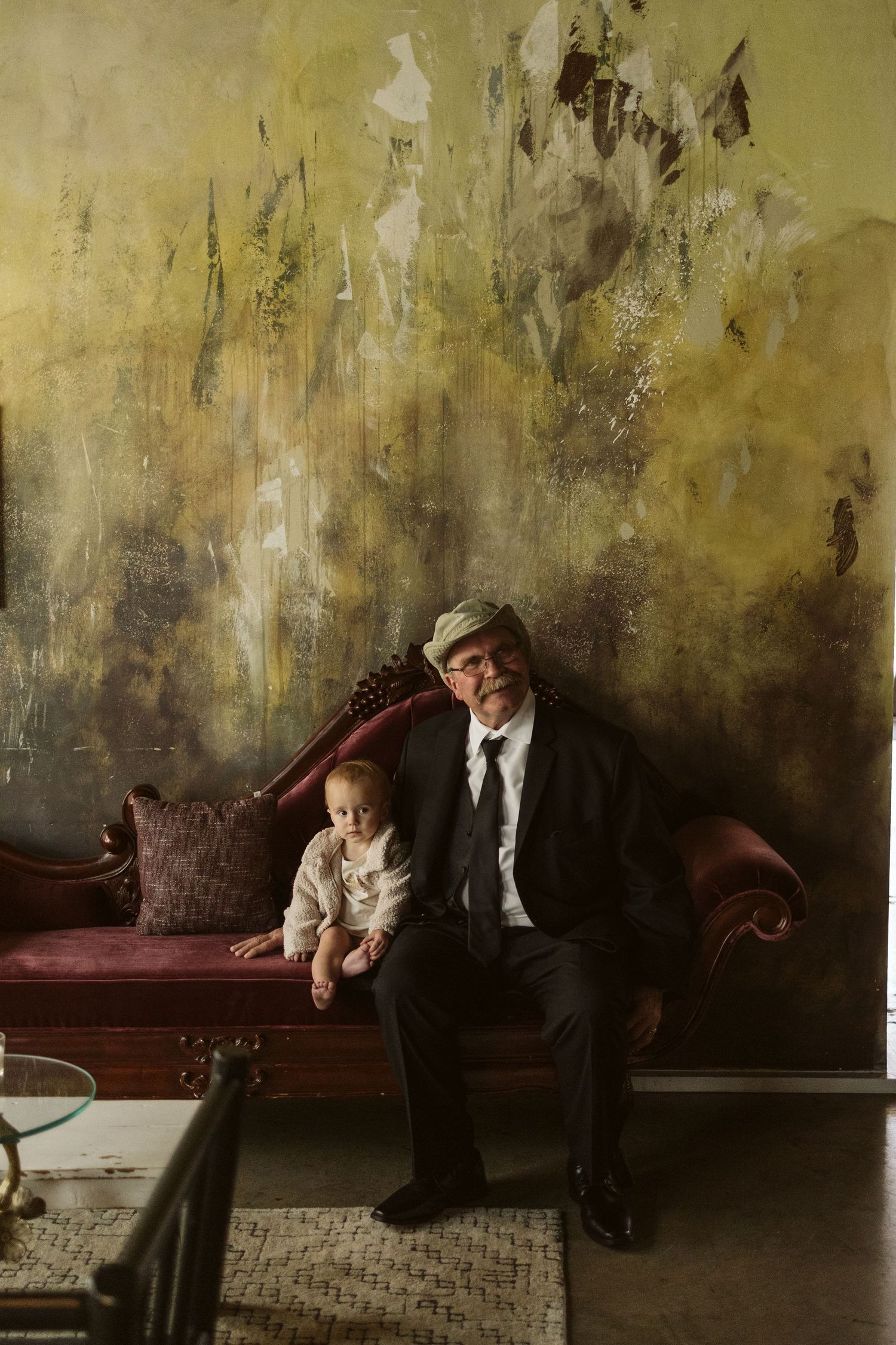 A vintage-style photo of two figures seated on an antique red velvet couch against a textured gold wall.