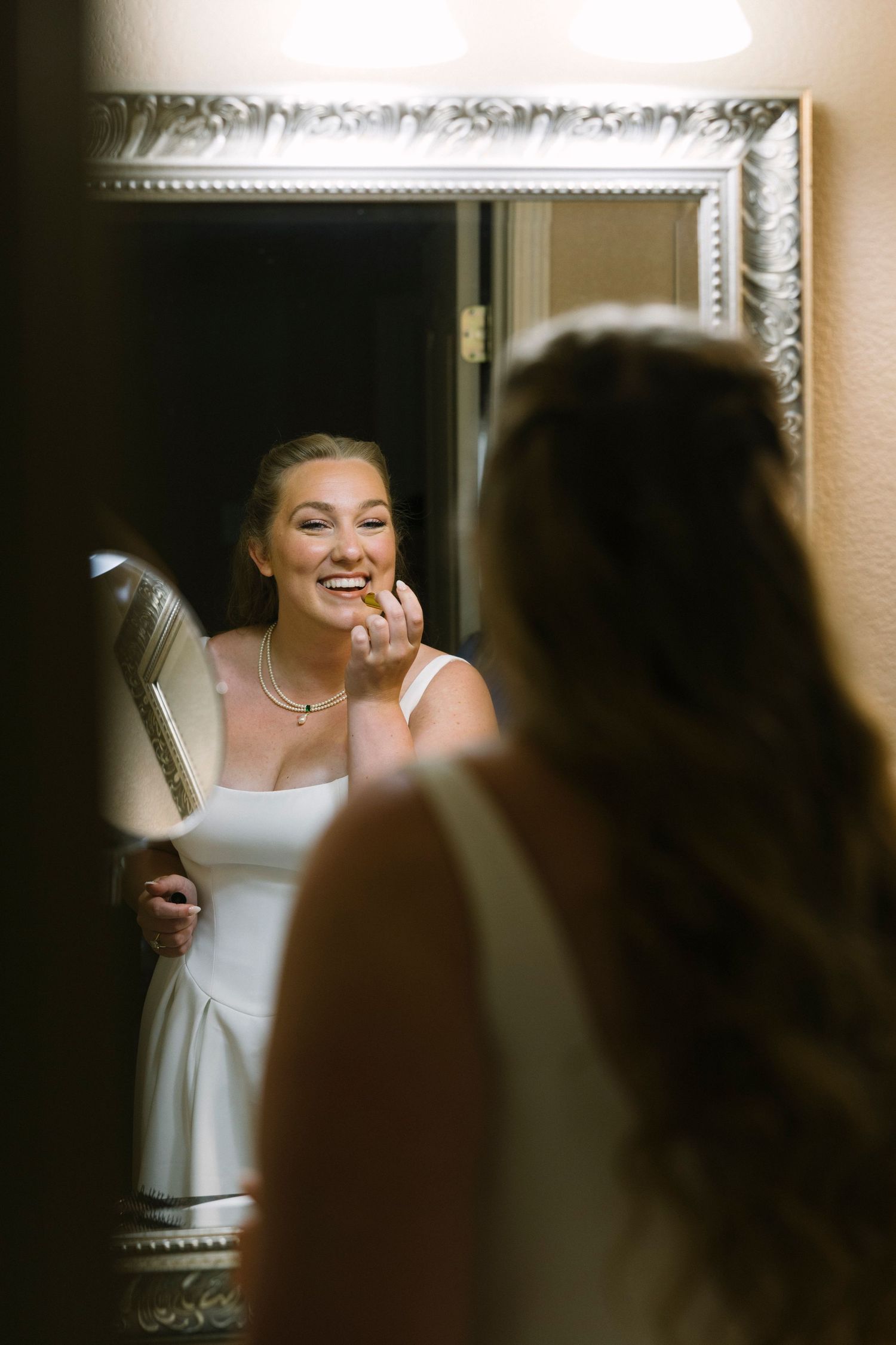 Reflection in ornate silver mirror shows joyful bride in white dress laughing while getting ready for wedding day.