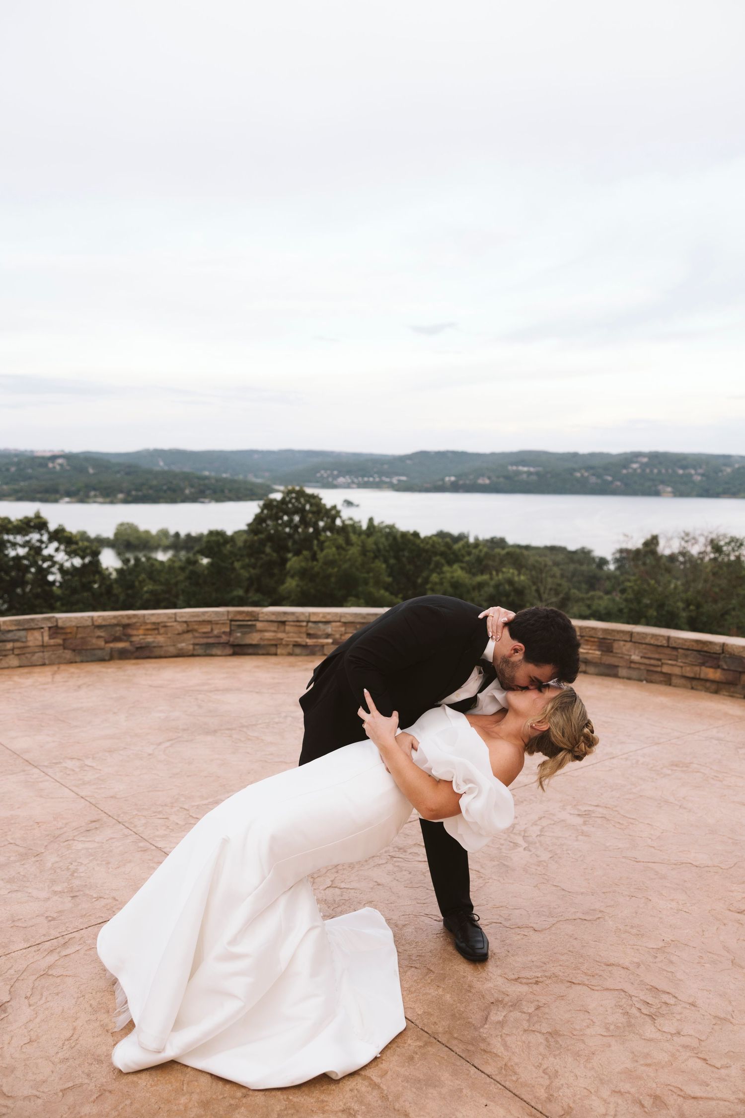 A couple shares a romantic dip-style dance pose on an outdoor terrace overlooking a scenic lake and rolling hills.