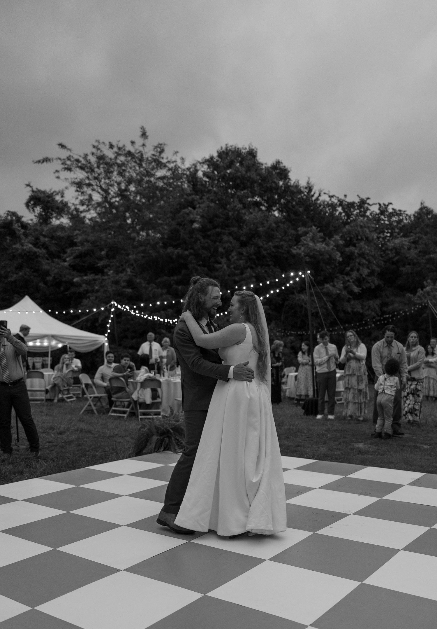 A couple shares their first dance on a black and white checkered dance floor at an outdoor wedding reception beneath string lights.