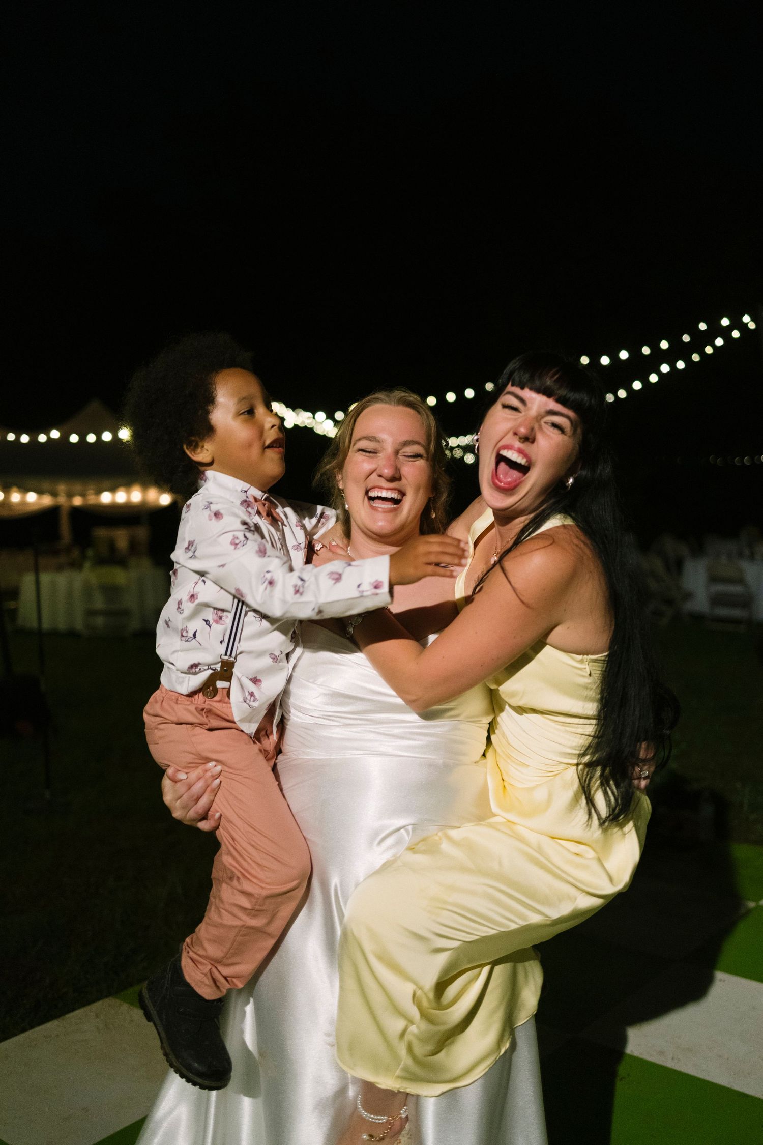 Group sharing joyful laughter and embraces while wearing light-colored dresses at an outdoor evening event under string lights.